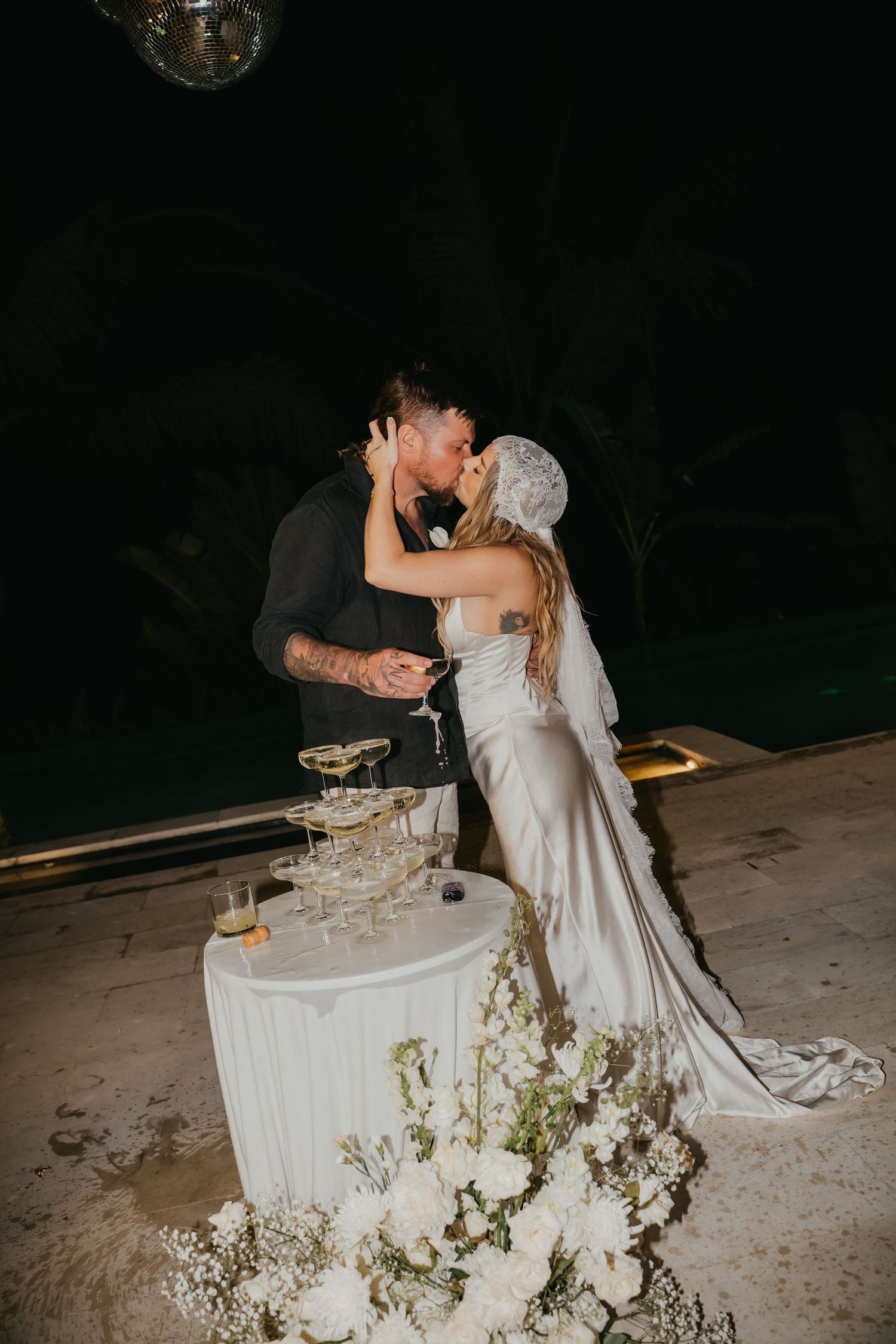 Newlyweds kiss beside champagne tower surrounded by white florals at romantic poolside night reception.