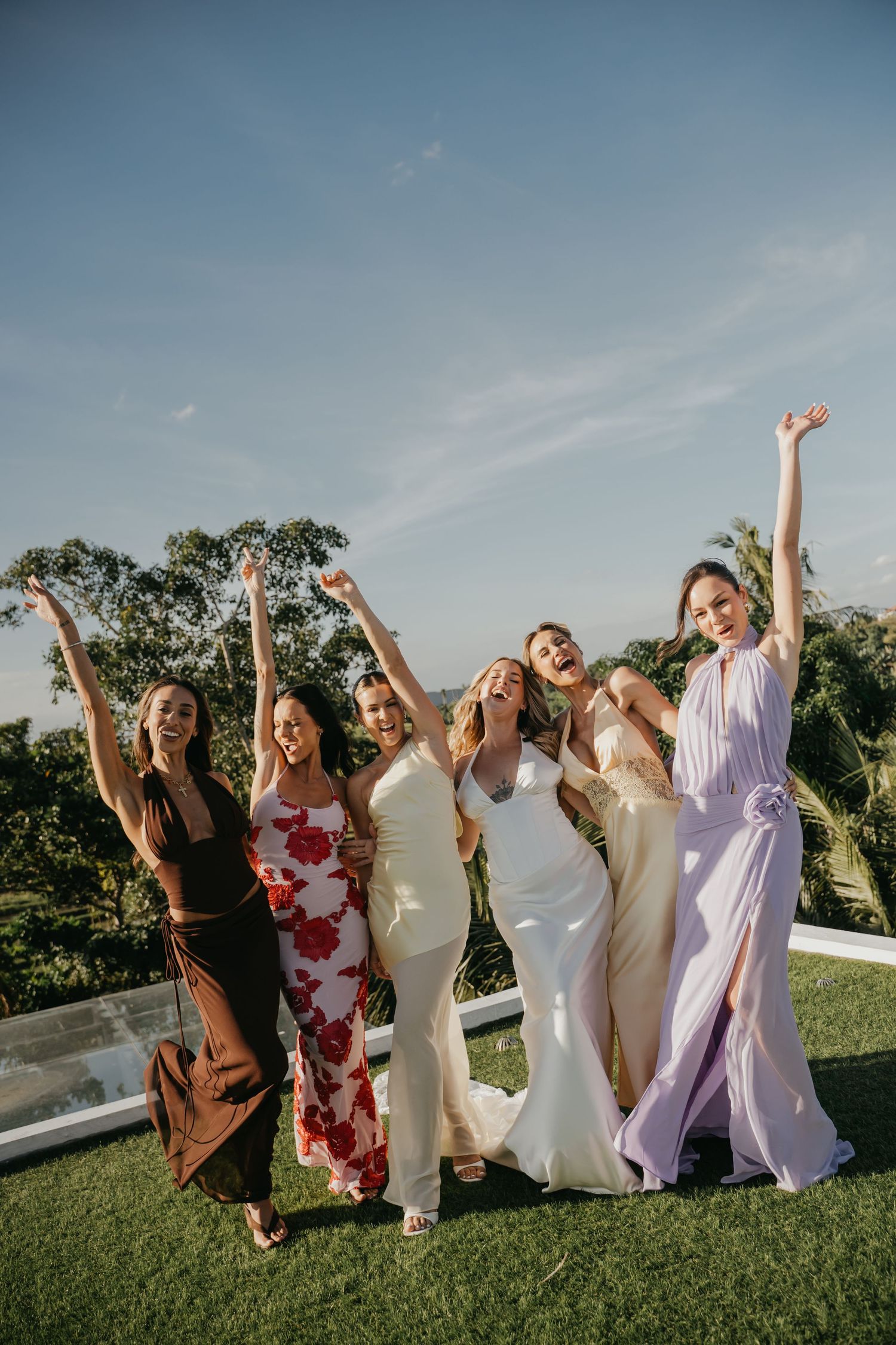 Joyful bride celebrates with bridesmaids in colorful dresses raising arms outdoors at wedding reception.
