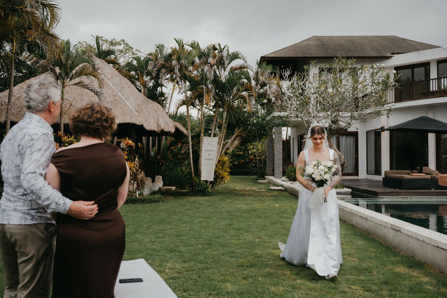 Bride in white gown walks toward family guests on lush green lawn of tropical villa wedding venue.