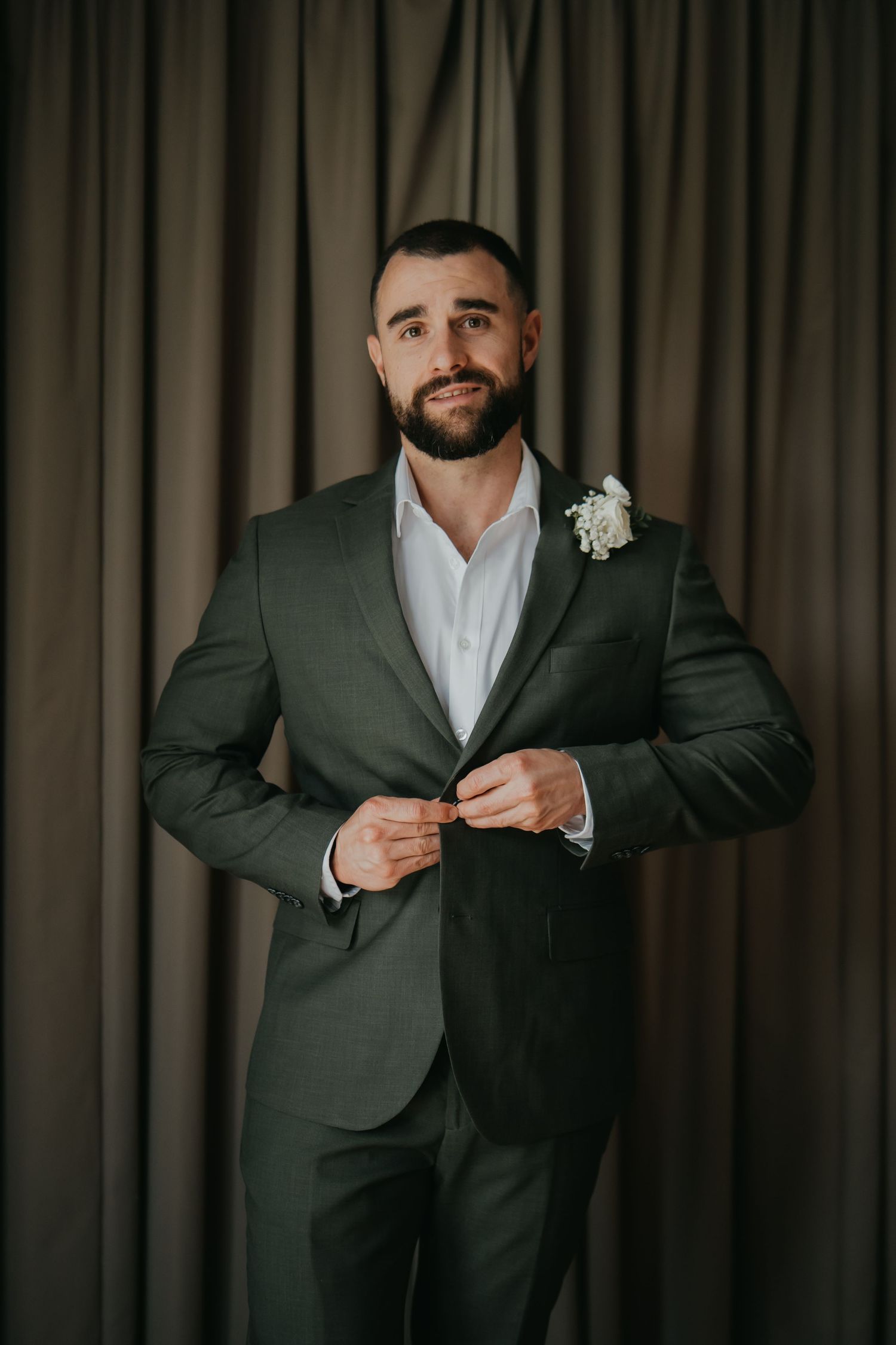 Groom in olive green suit with white boutonniere buttoning jacket before wedding ceremony.