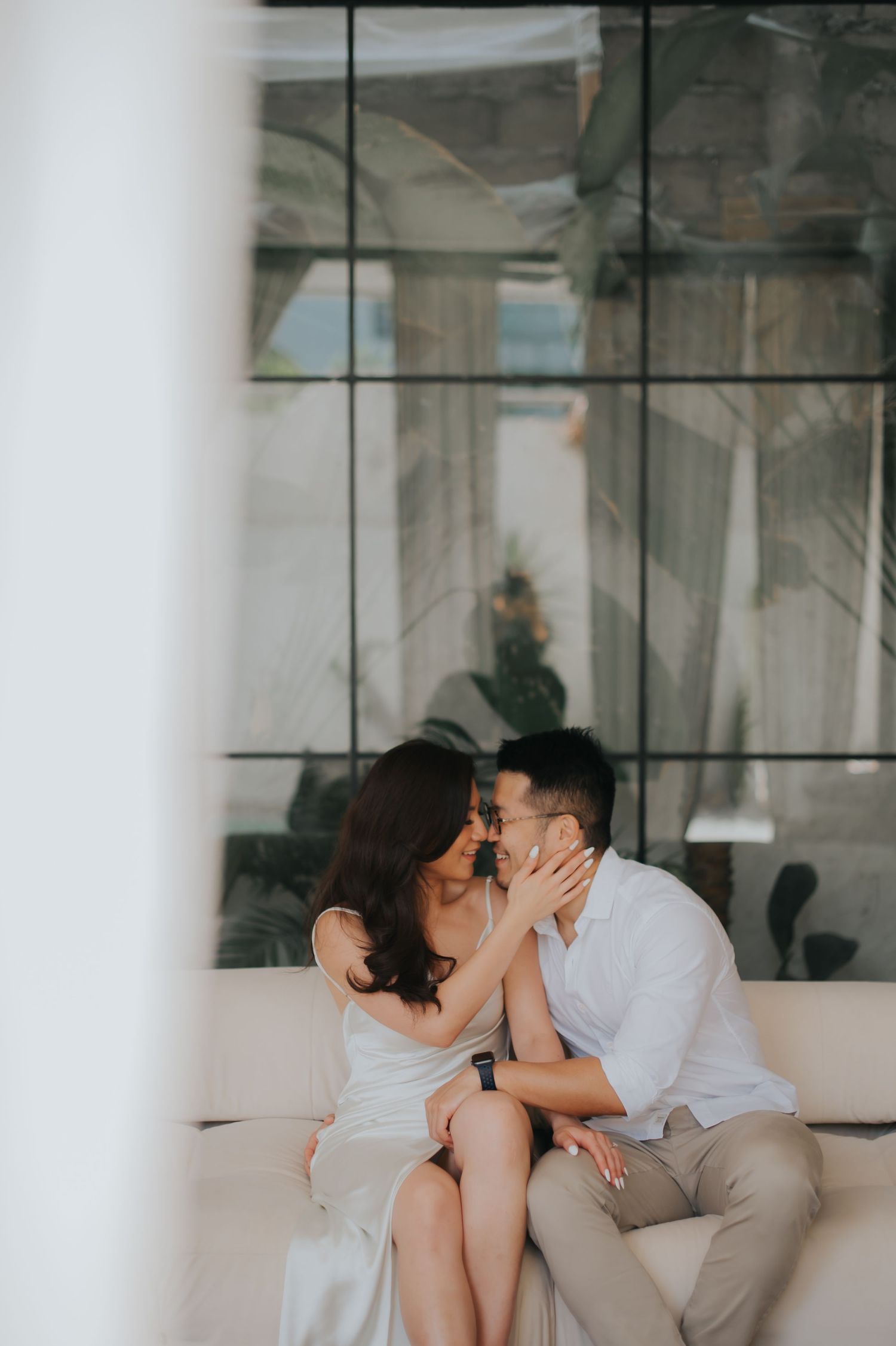 A couple shares a romantic moment on a white sofa, with the woman gently touching the man's face in an elegant indoor setting.