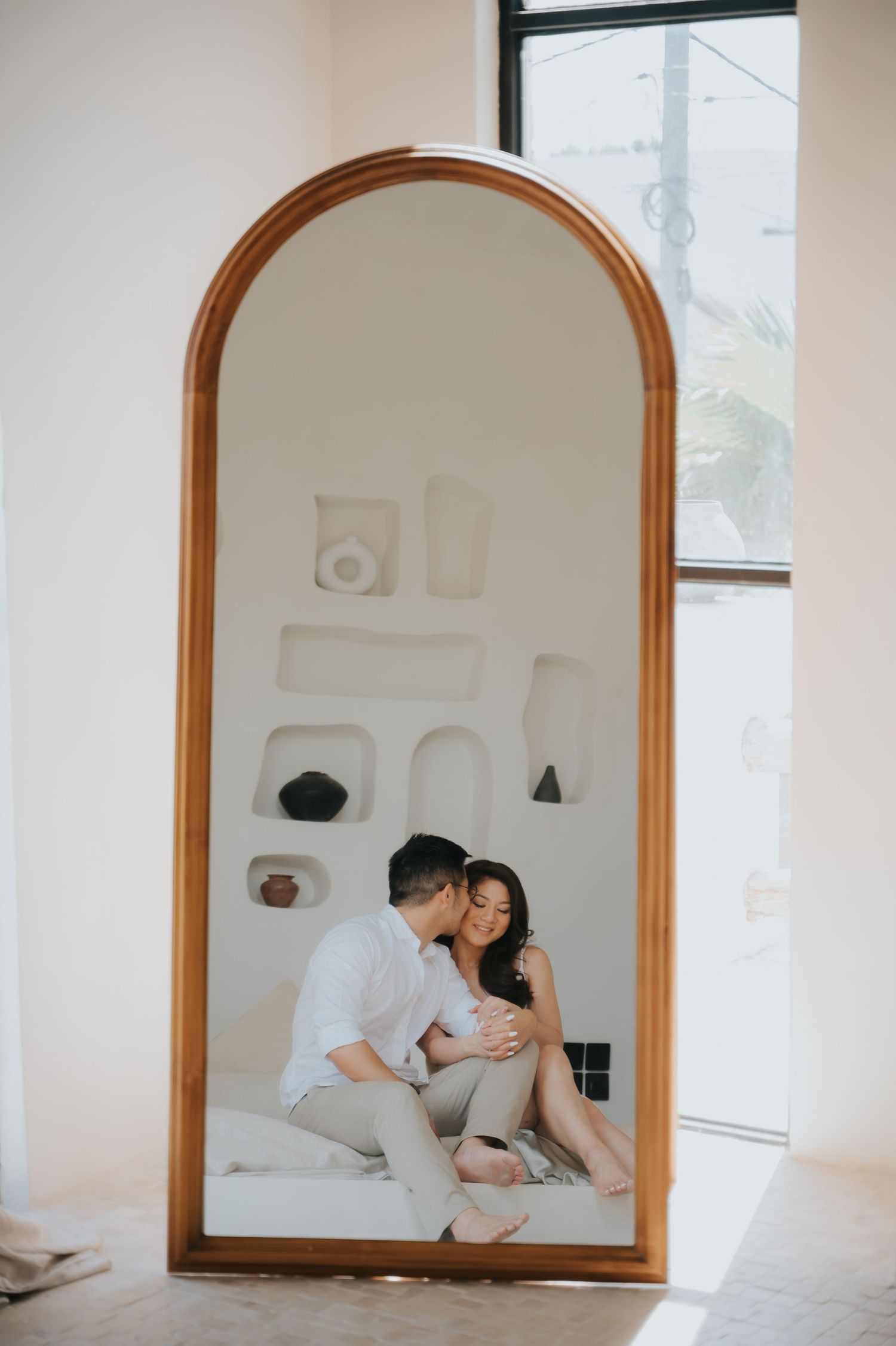 Couple sharing an intimate moment reflected in a large arched wooden mirror in a bright, minimalist white room.