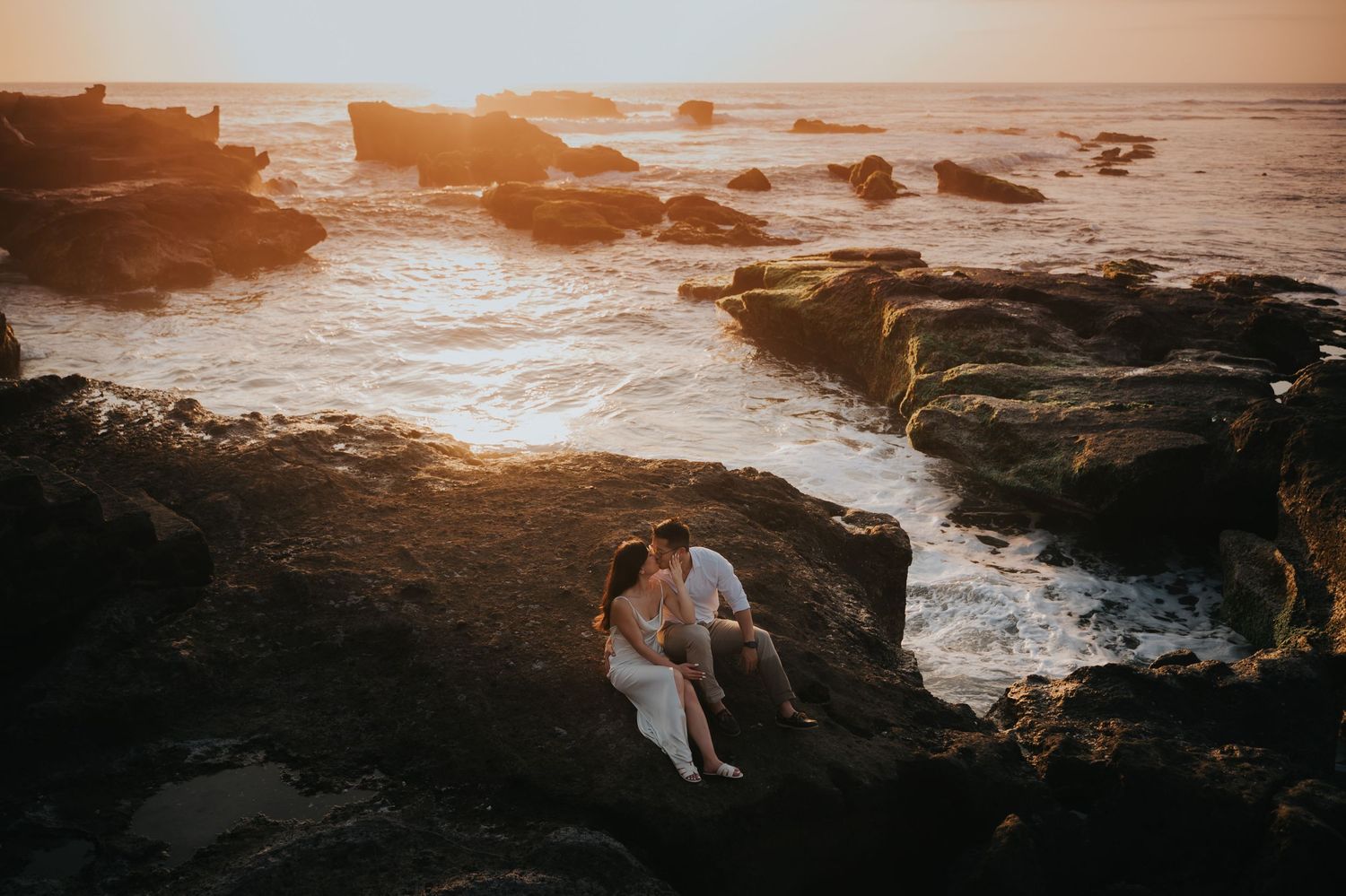 Couple sitting close together on rocky coastal cliffs at golden sunset with ocean waves crashing around them.