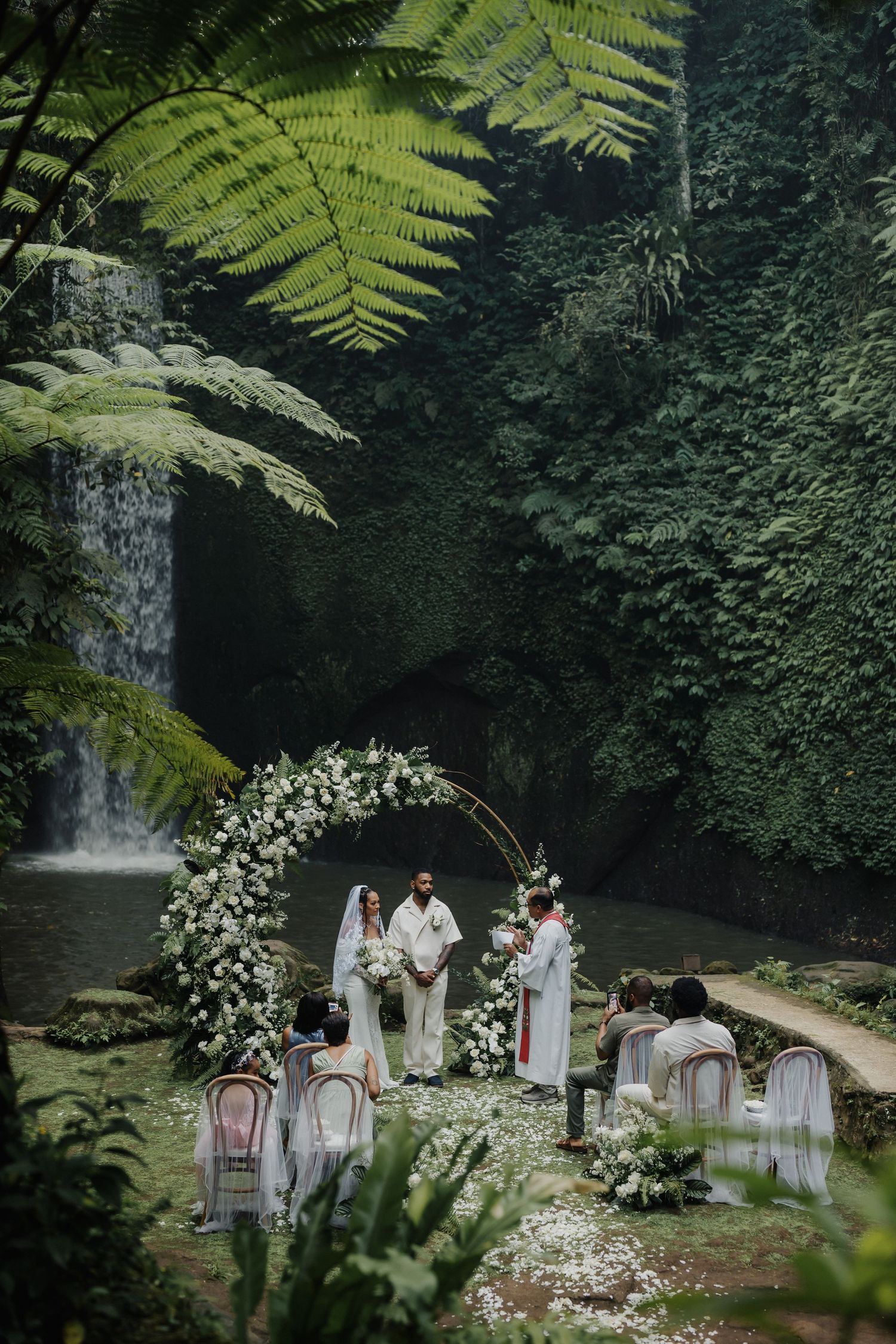Intimate outdoor wedding ceremony beside a tropical waterfall with white floral arch and lush greenery.