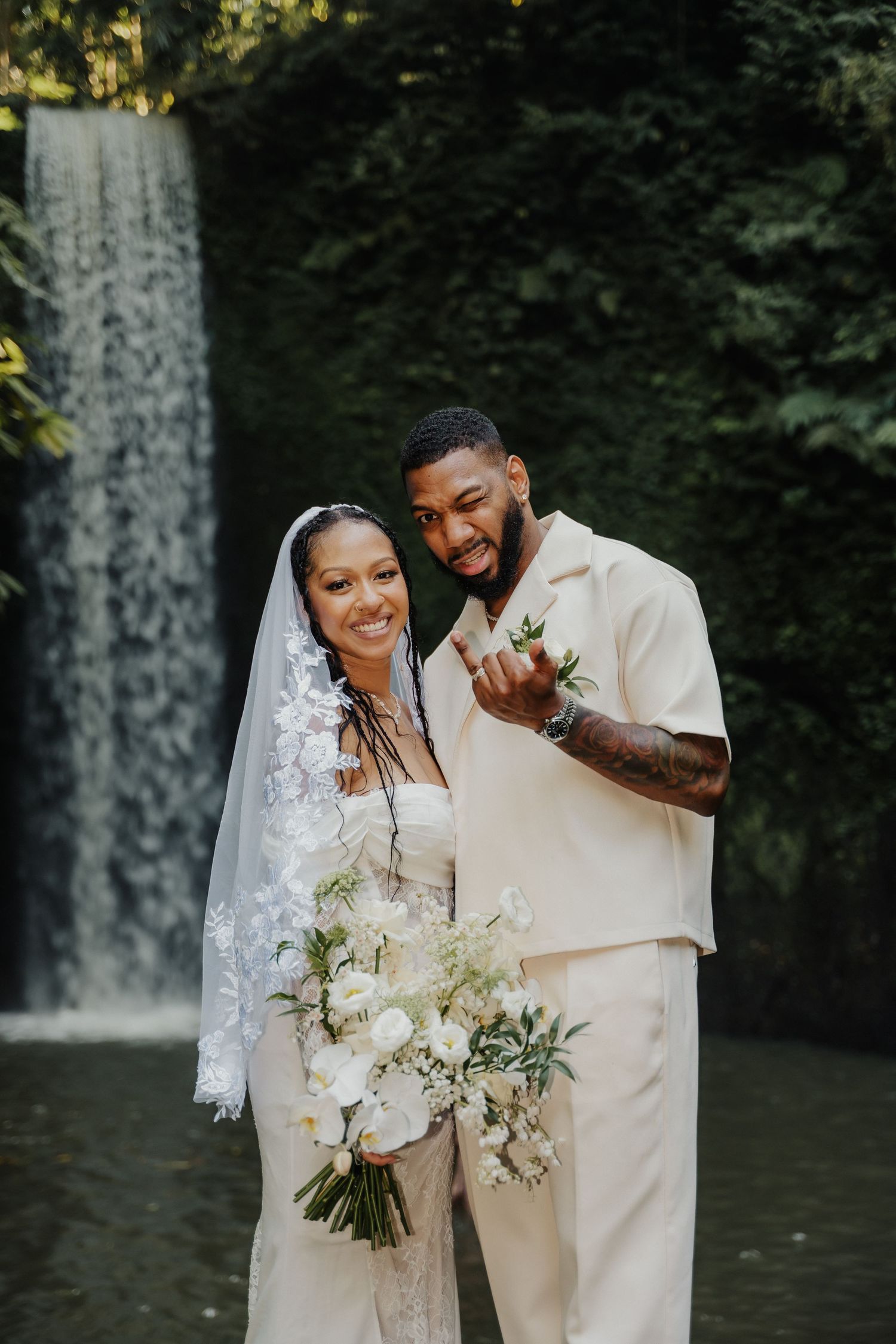 A smiling couple in cream wedding attire poses in front of a lush waterfall, bride holding white floral bouquet.