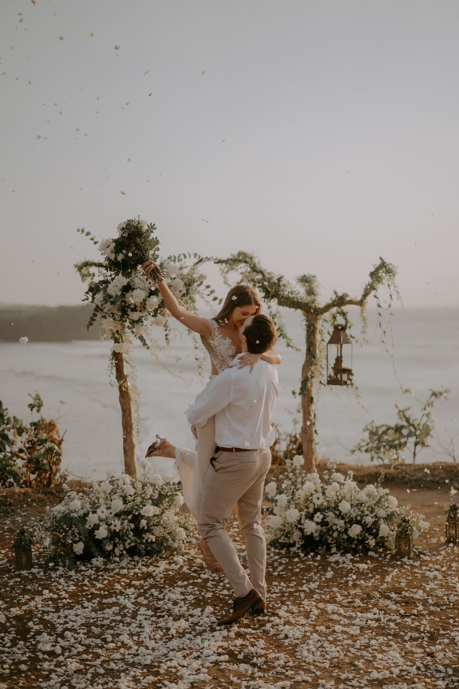 A couple dances together on a cliff overlooking the ocean at sunset, surrounded by scattered flower petals.