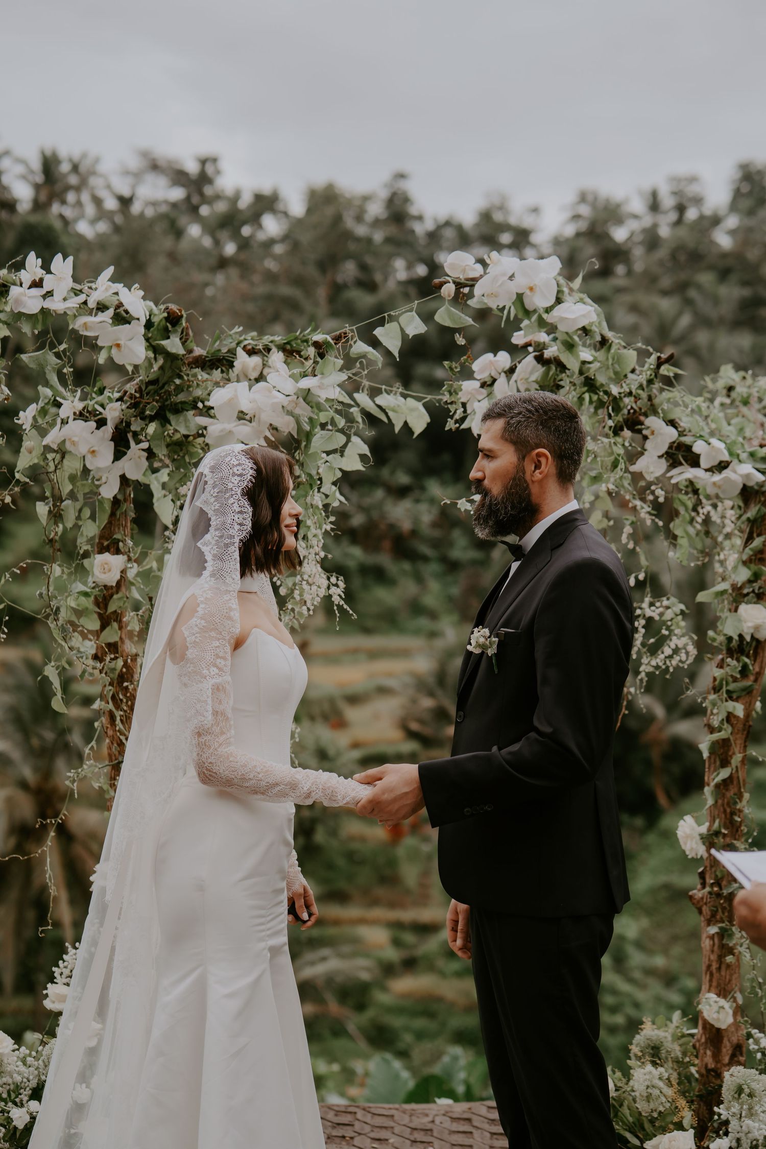 A romantic outdoor wedding ceremony under a floral arch with white blooms and greenery in a garden setting.