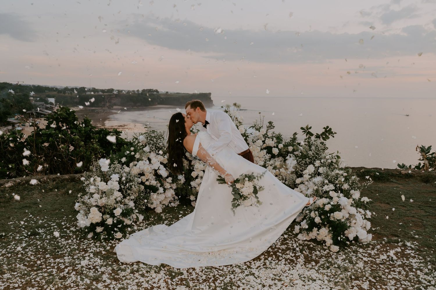 Bride and groom share a romantic dip kiss among white floral arrangements at sunset.