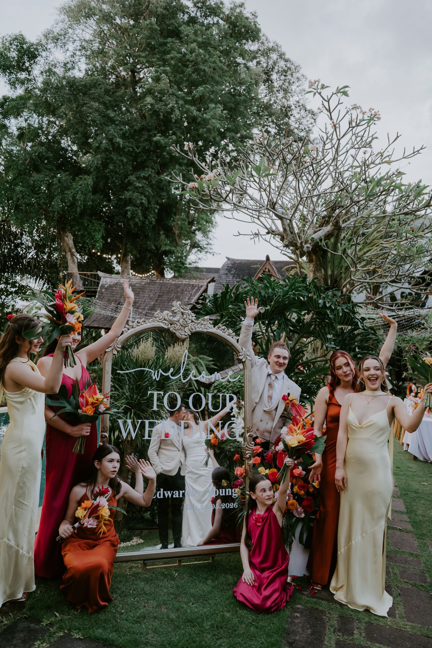 Bride, groom, and bridesmaids in colorful dresses posing by a Welcome to Our Wedding mirror sign outdoors.