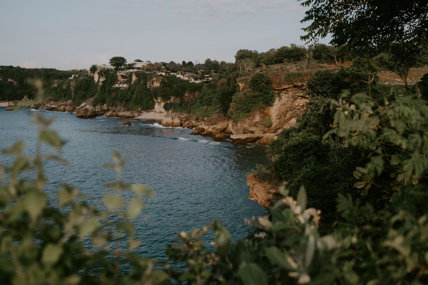 Rocky coastal cliffs covered in lush greenery overlook a calm blue bay in a scenic Mediterranean landscape.