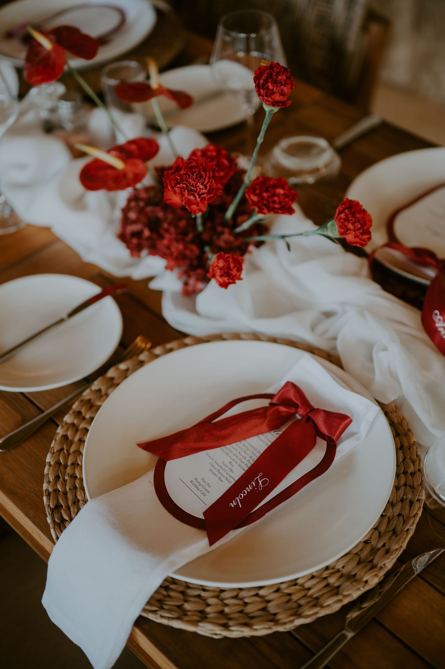 Elegant wedding table setting with red floral centerpieces, white plates, and ribbon-tied menus.