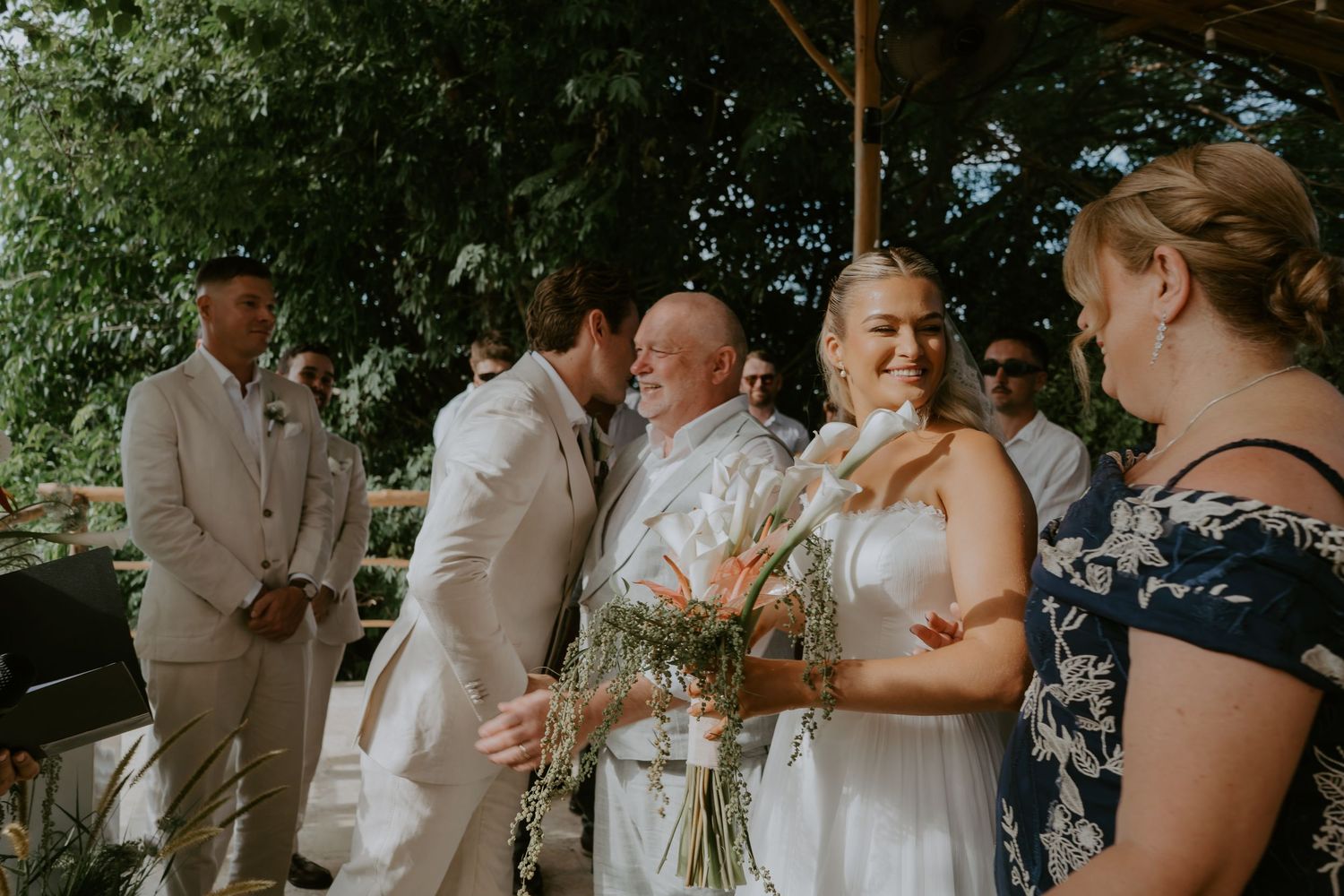 Bride smiling with bouquet receives a kiss from groom during an outdoor tropical wedding ceremony.