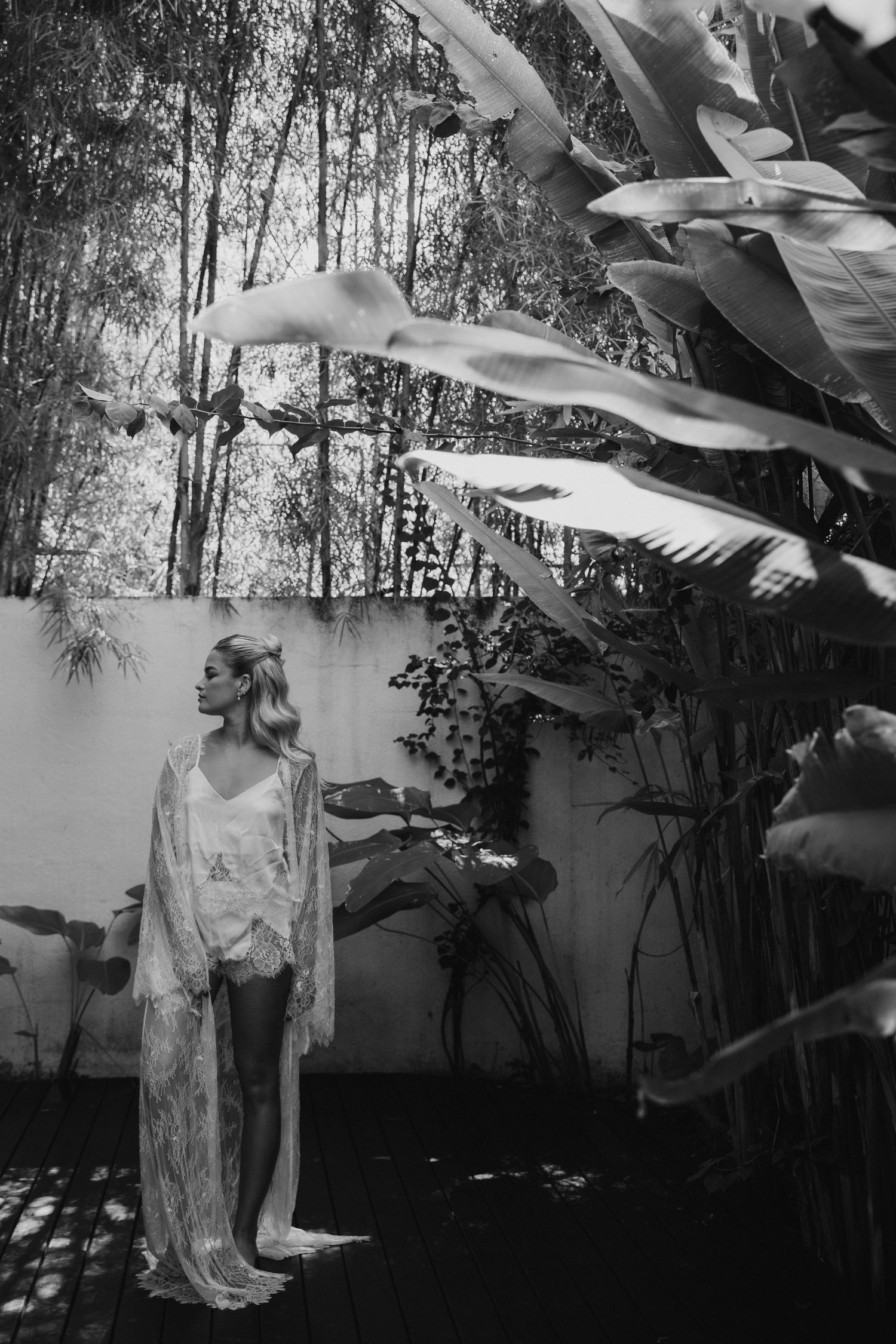 Bride in elegant lace robe standing among tropical plants in black and white wedding portrait.