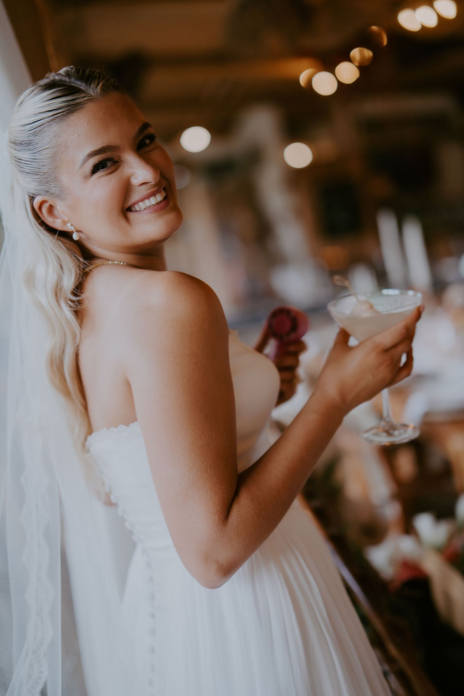 Smiling bride in strapless white gown and veil holding a cocktail glass at her wedding reception.