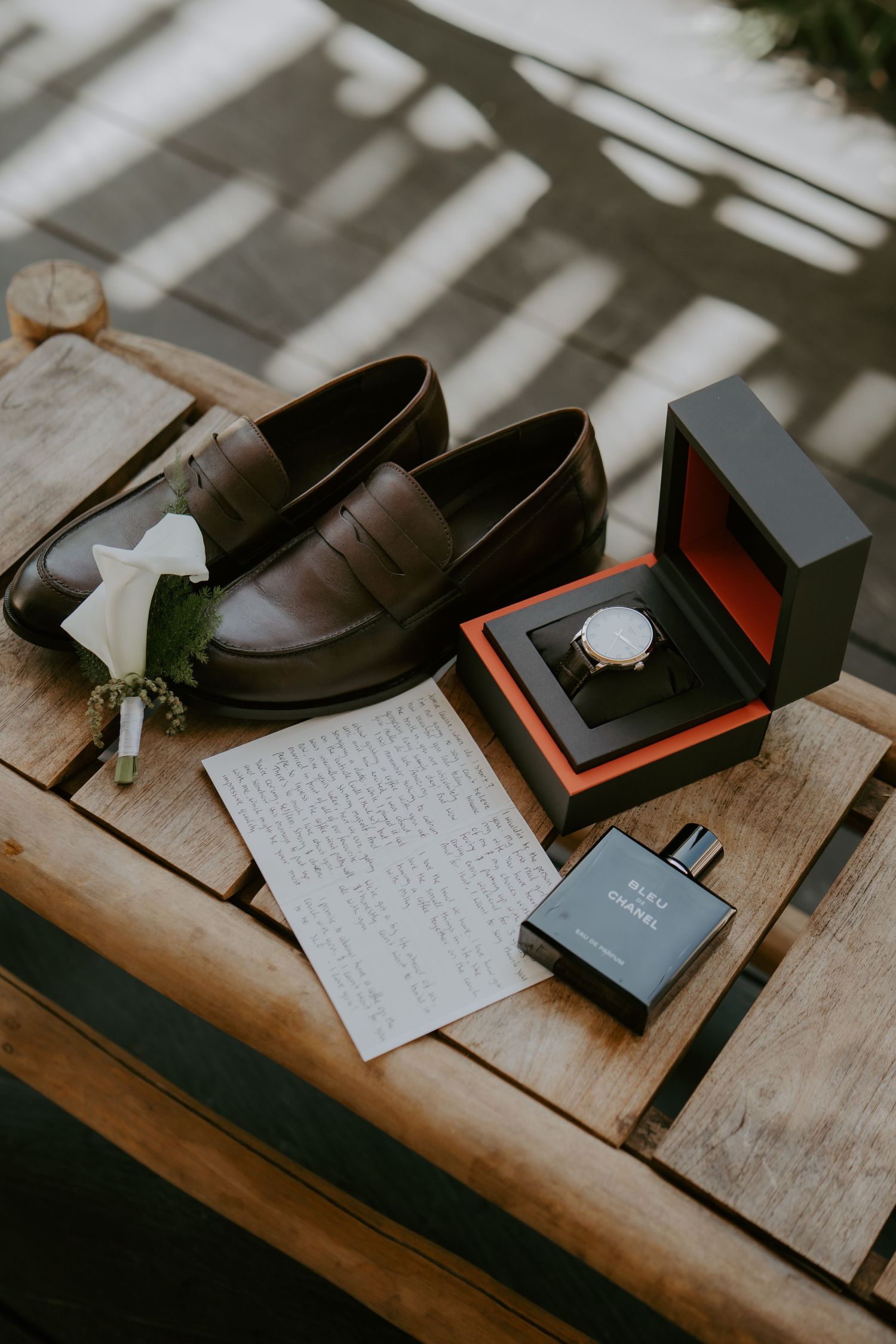 Groom's wedding accessories flat lay featuring brown loafers, watch in box, boutonniere, handwritten note, and Chanel cologne.