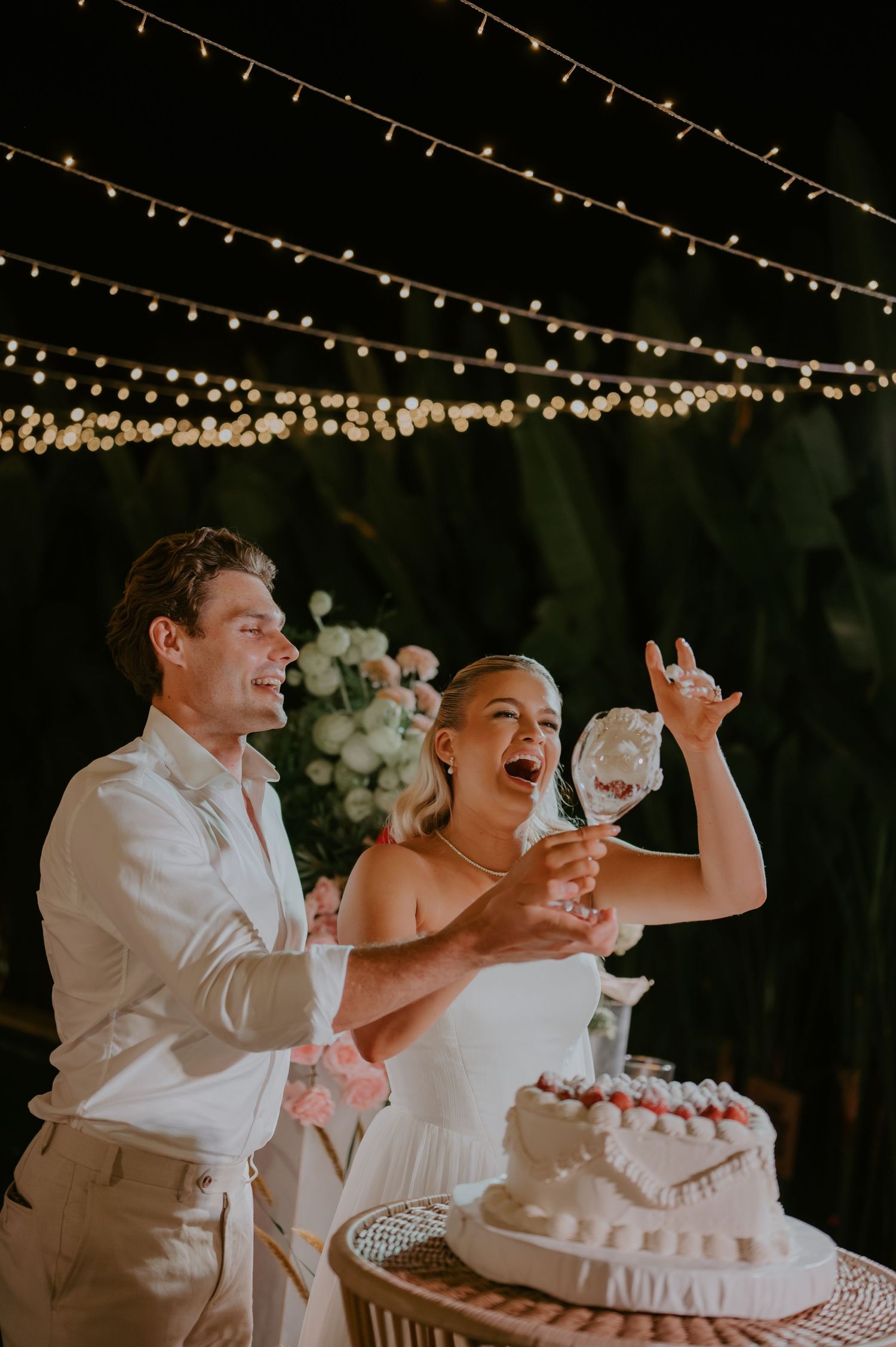Bride and groom laughing while cutting wedding cake under string lights at outdoor night reception.