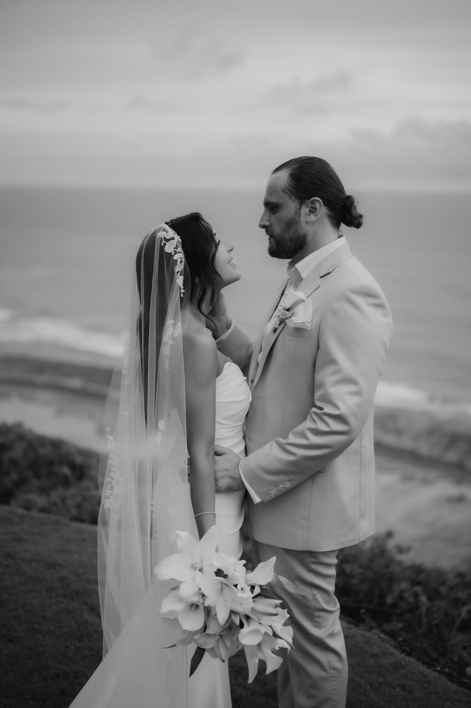 Black and white portrait of bride and groom gazing at each other with ocean view behind them.