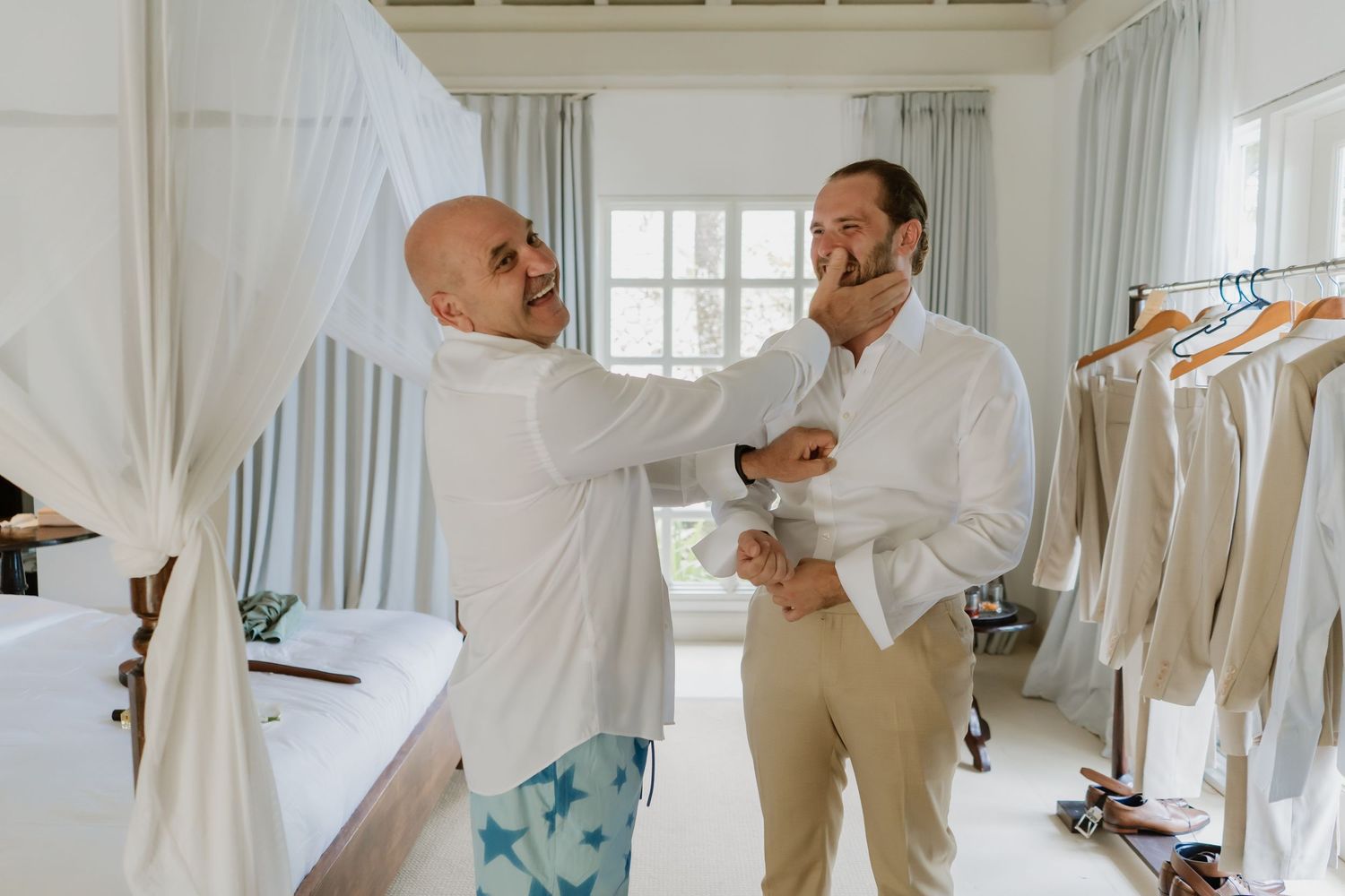 Two men in a bright room, one adjusting the other's shirt collar, preparing for a wedding ceremony.