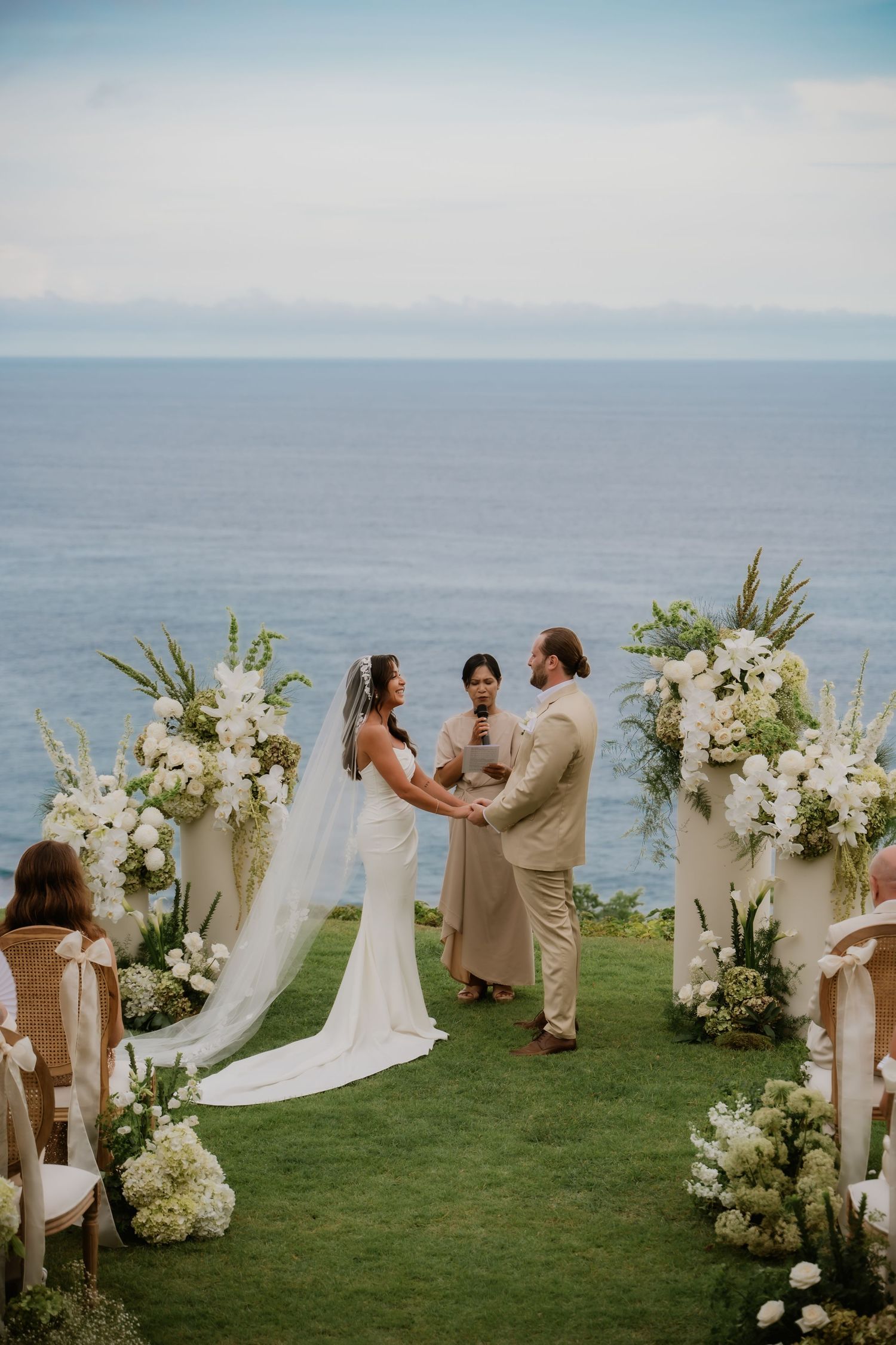 Bride and groom exchange vows at oceanfront ceremony with white floral arrangements.