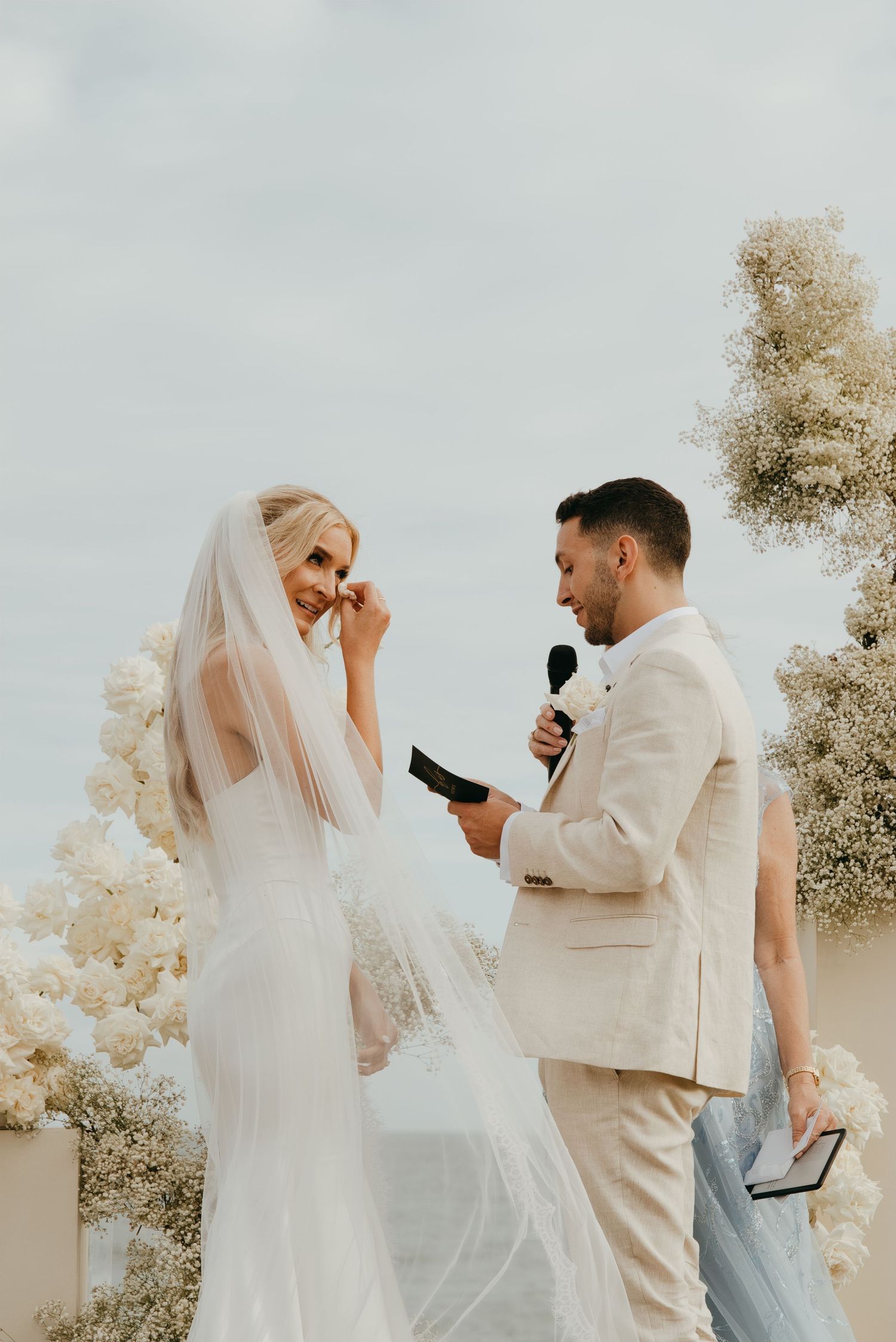 Emotional bride in white gown and veil wipes tears as groom in beige suit reads wedding vows at outdoor ceremony.