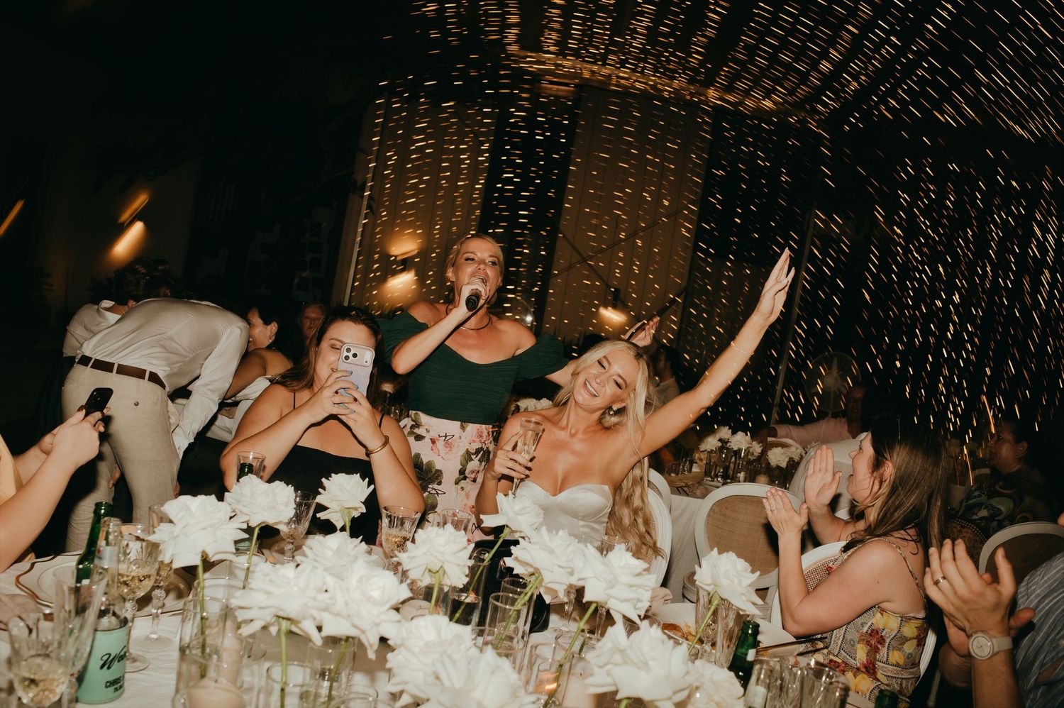 Bride and guests joyfully singing and celebrating at a fairy-light adorned wedding reception dinner.