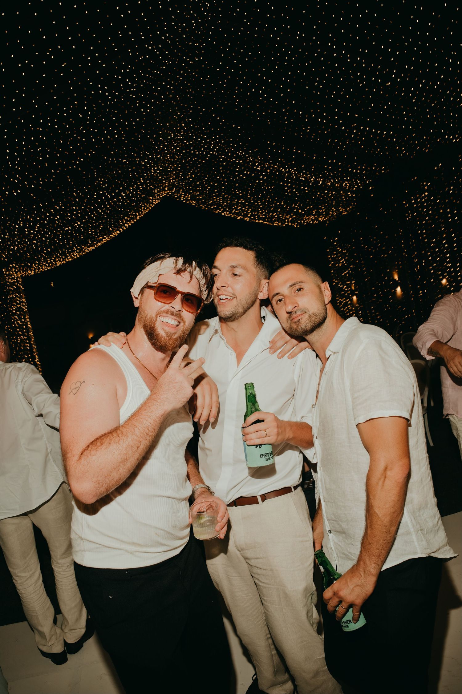 Three male wedding guests posing together under fairy lights while holding beer bottles at reception.