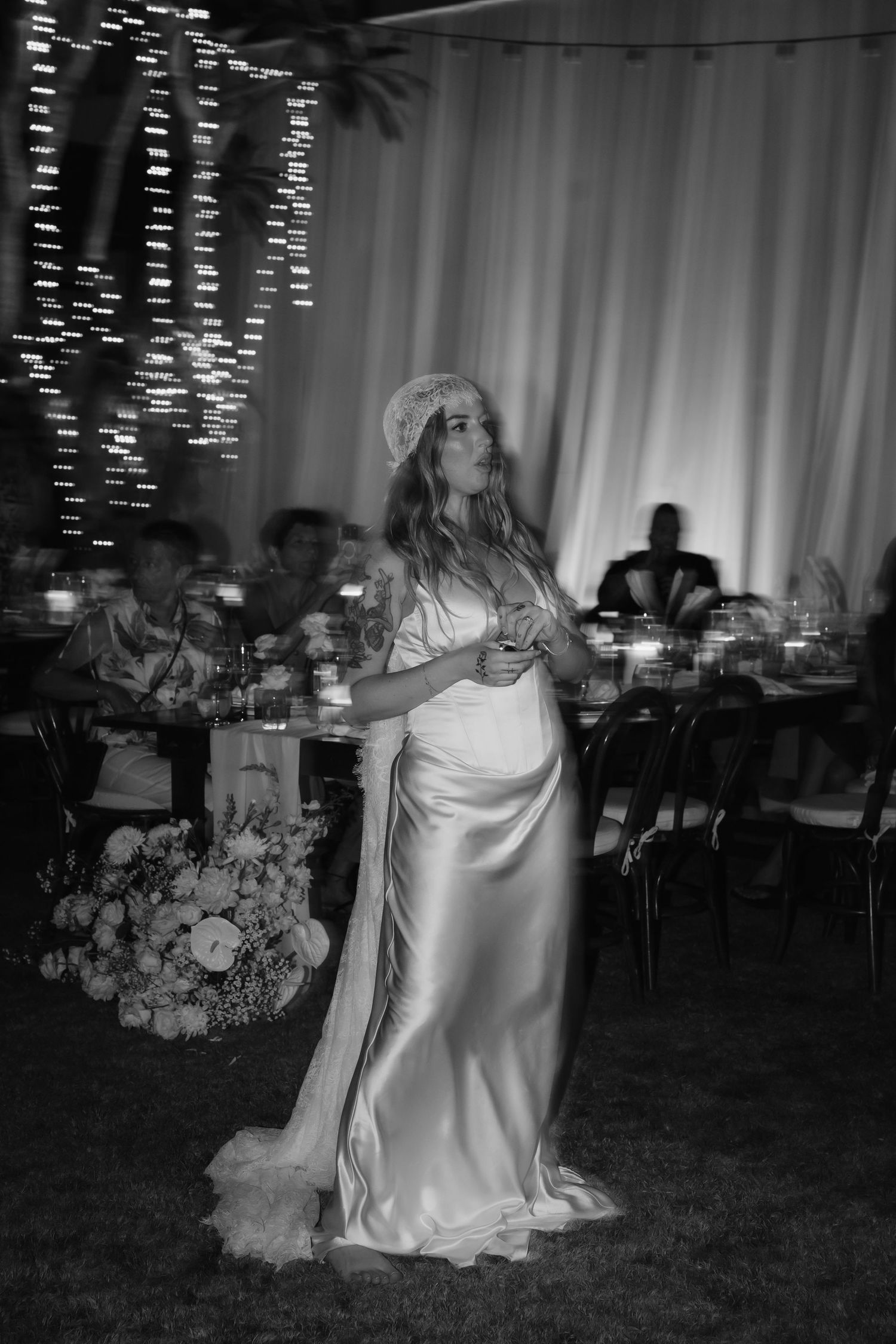 Black and white photo of bride in satin gown and lace cap holding flowers at candlelit reception.