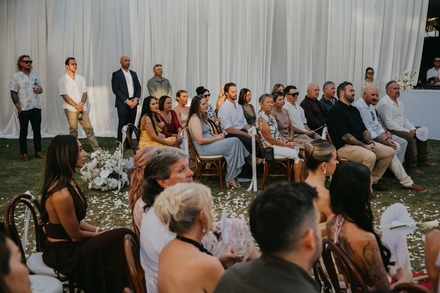 Wedding guests seated in wooden chairs at outdoor tropical ceremony with white draping and lush greenery backdrop.