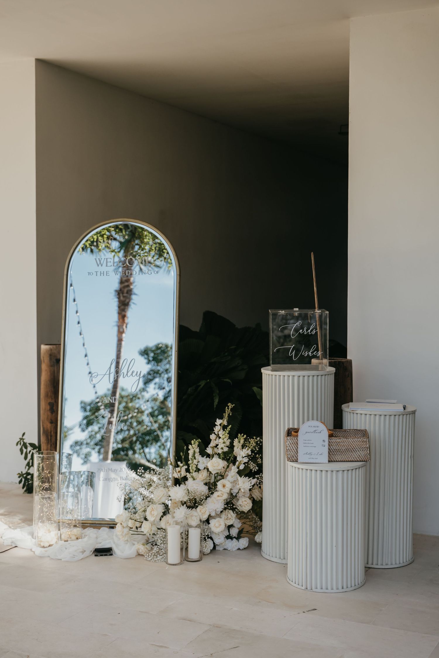 Elegant wedding welcome display with arch mirror, white floral arrangements, and decorative pedestals.