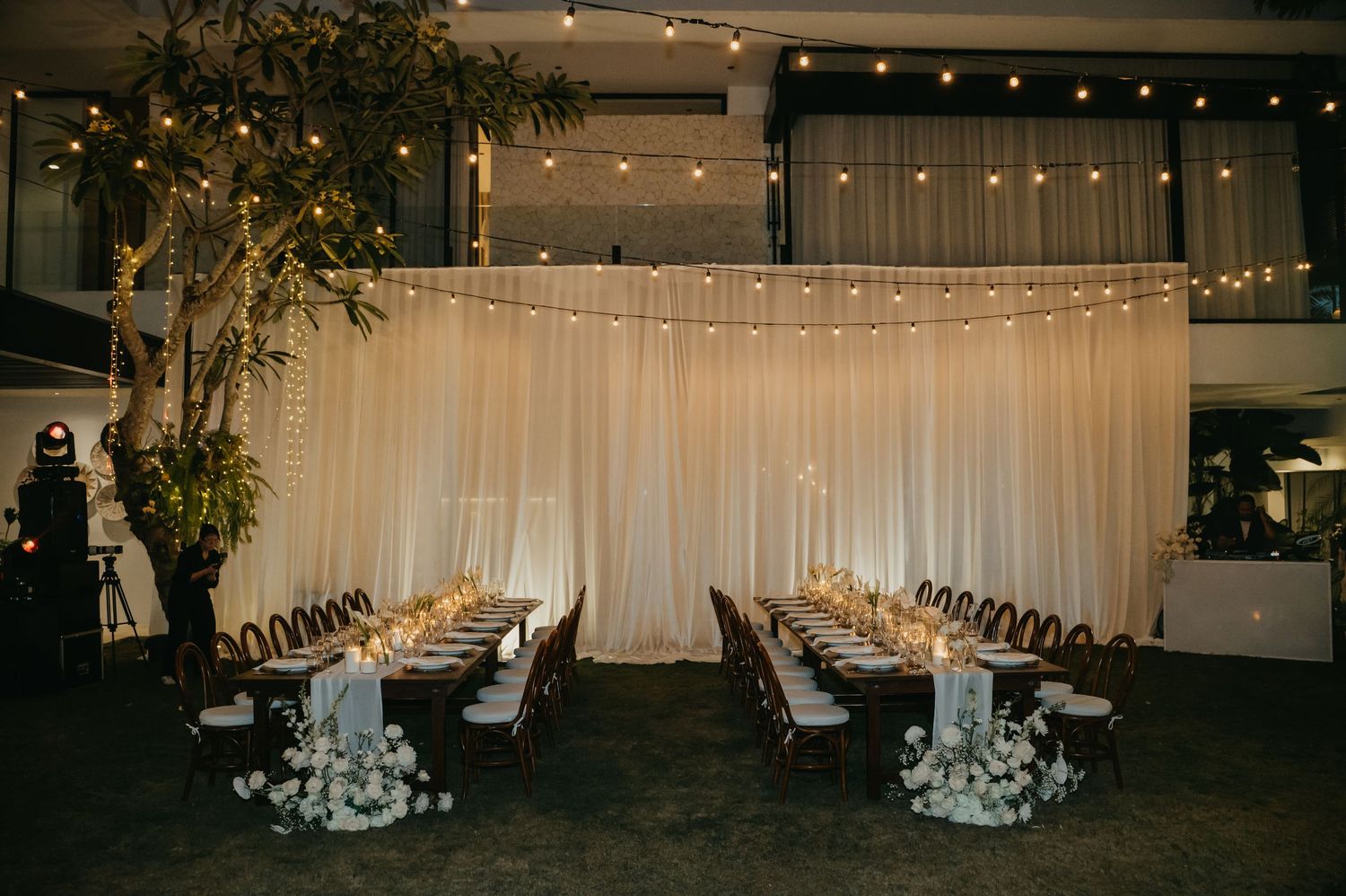 Elegant indoor wedding reception with long tables, candlelight, white floral centerpieces, and string lights against draped backdrop.