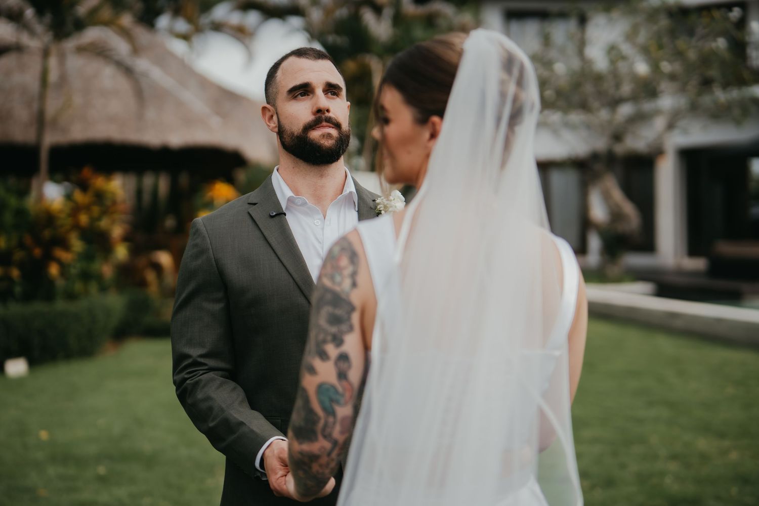 Tattooed groom in grey suit holding bride's hand outdoors at garden wedding ceremony.