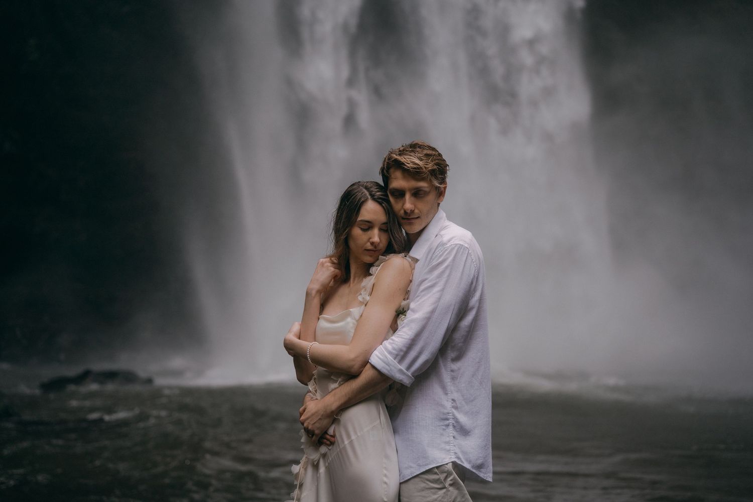A romantic couple embraces near a misty waterfall in a moody, atmospheric setting.