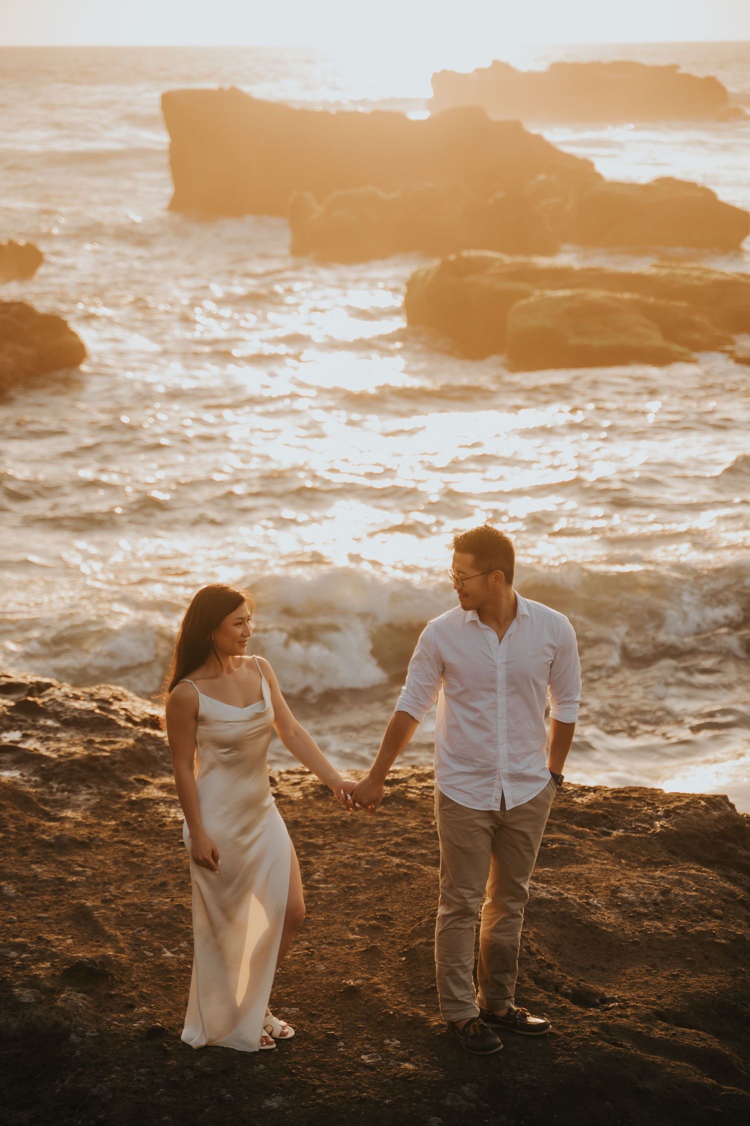 Couple holding hands on rocky ocean cliffs at golden sunset, woman in white dress, man in white shirt.