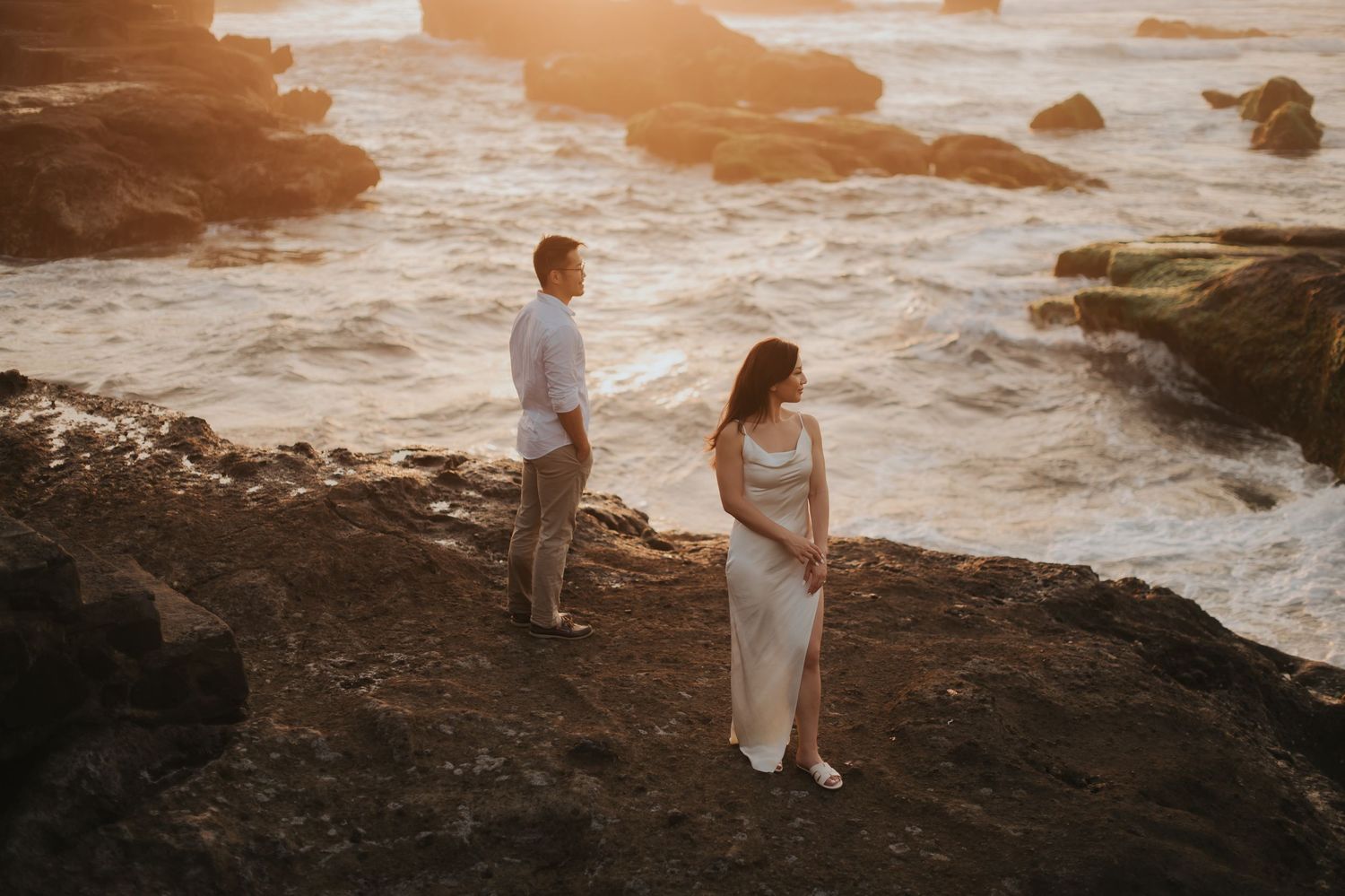 Couple standing on rocky coastal cliffs at golden sunset, waves crashing behind them, woman in white dress, man in white shirt.