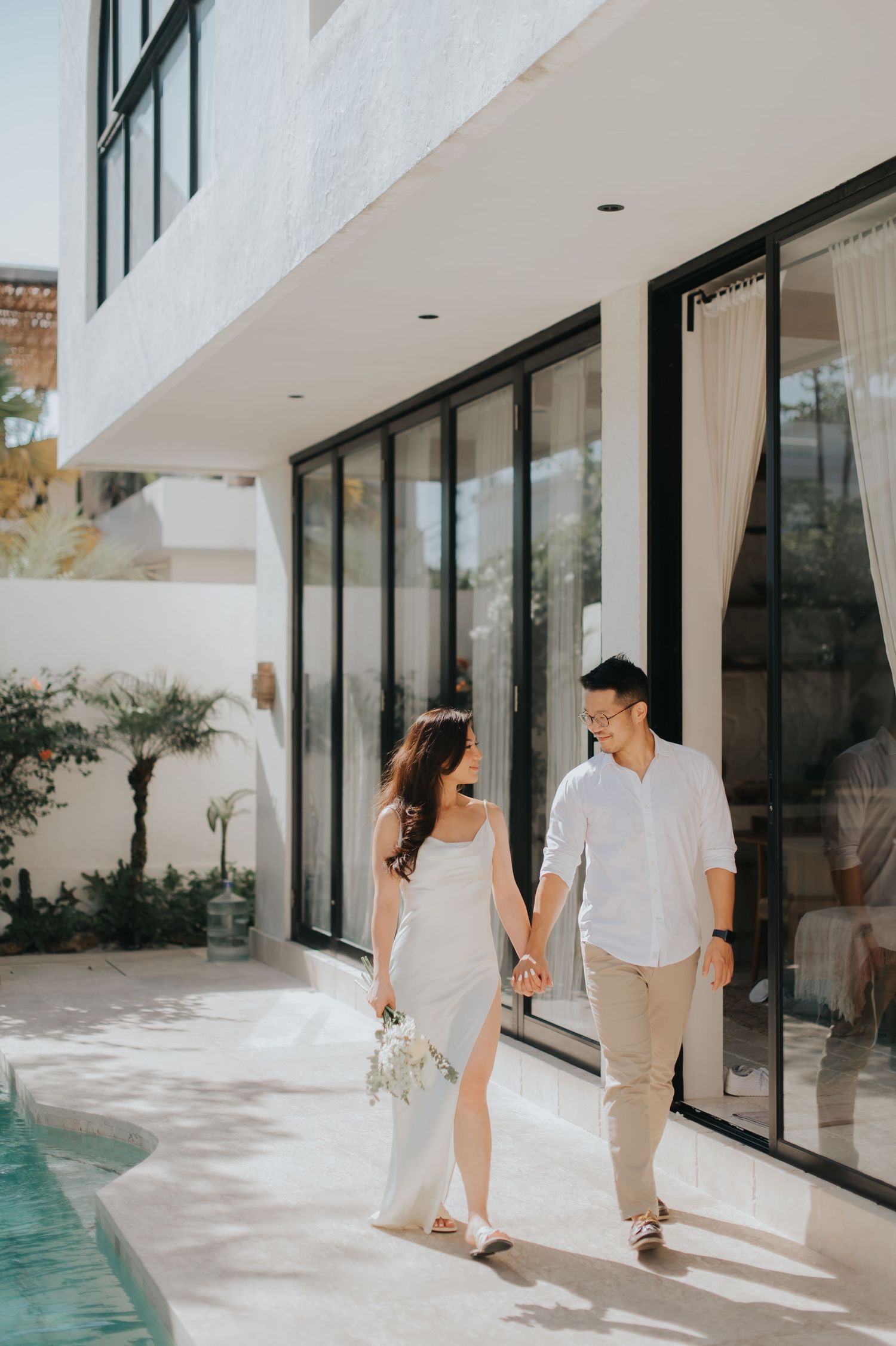 Couple in white outfits holding hands walking by a modern villa pool, woman holding a bouquet.