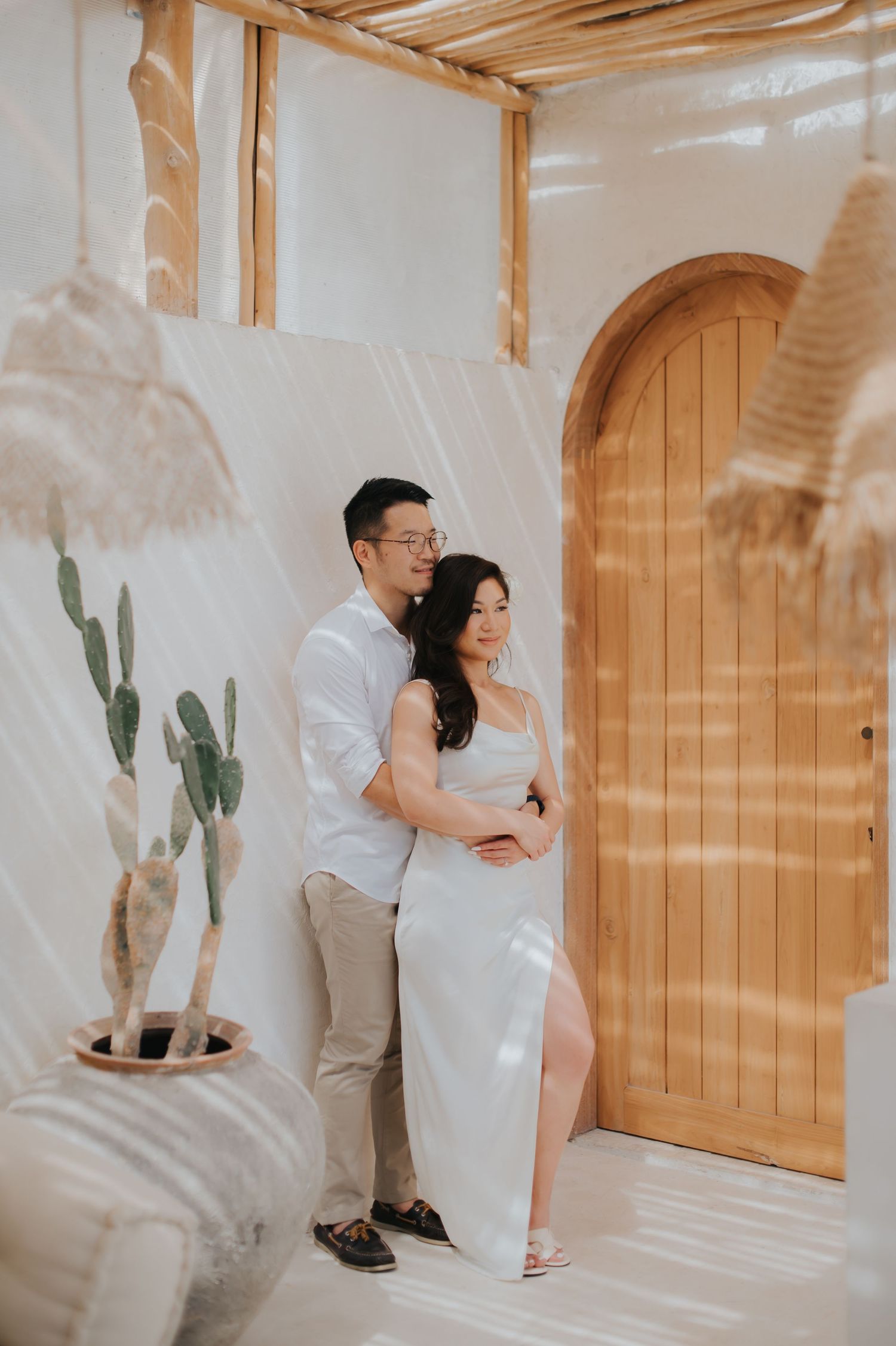 Couple in white outfits embracing in a boho-styled space with arched wooden door, cactus, and sheer white curtains.