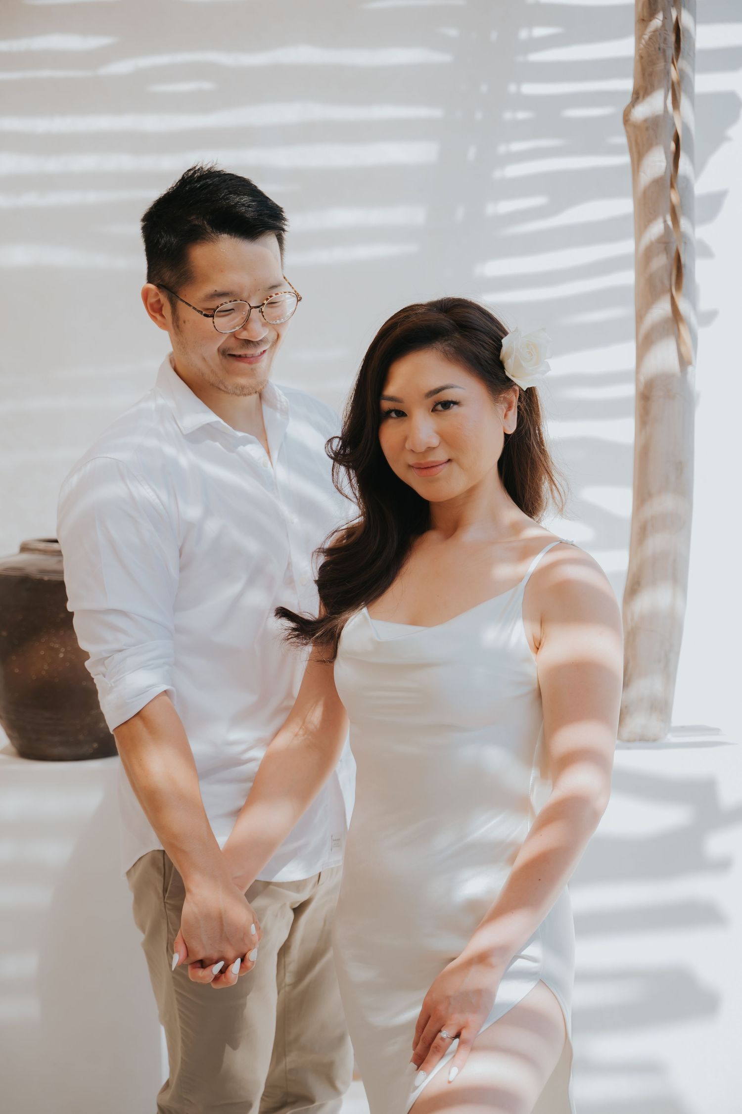 A couple in white outfits hold hands, the woman wearing a flower in her hair, posing elegantly in soft natural light.