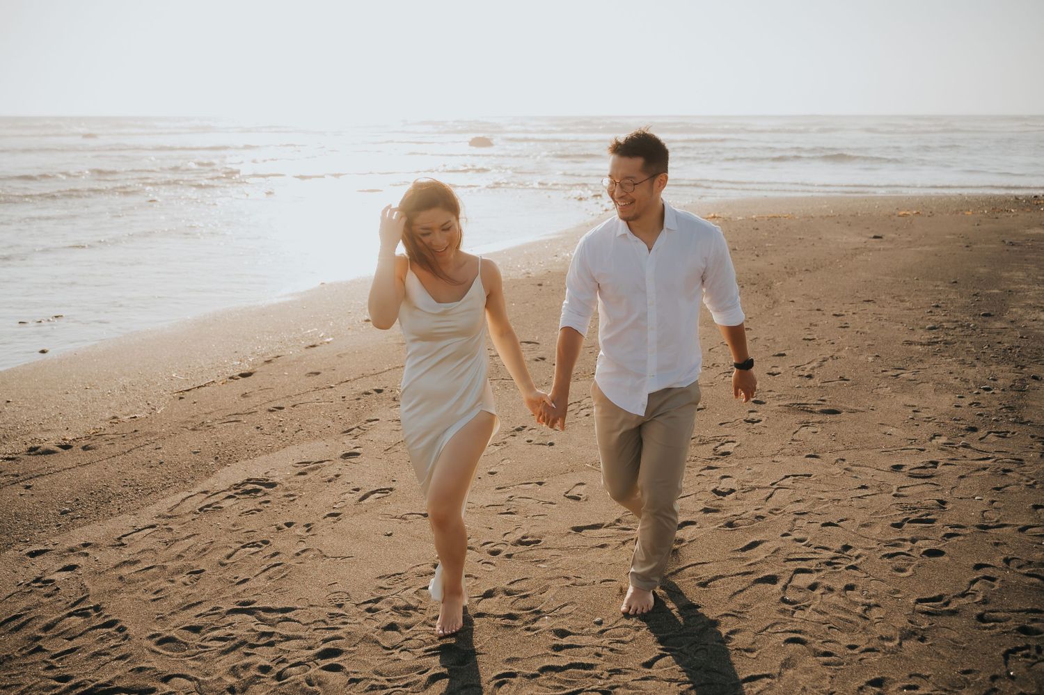A couple holds hands and walks along a sandy beach at sunset, smiling and enjoying a romantic stroll.