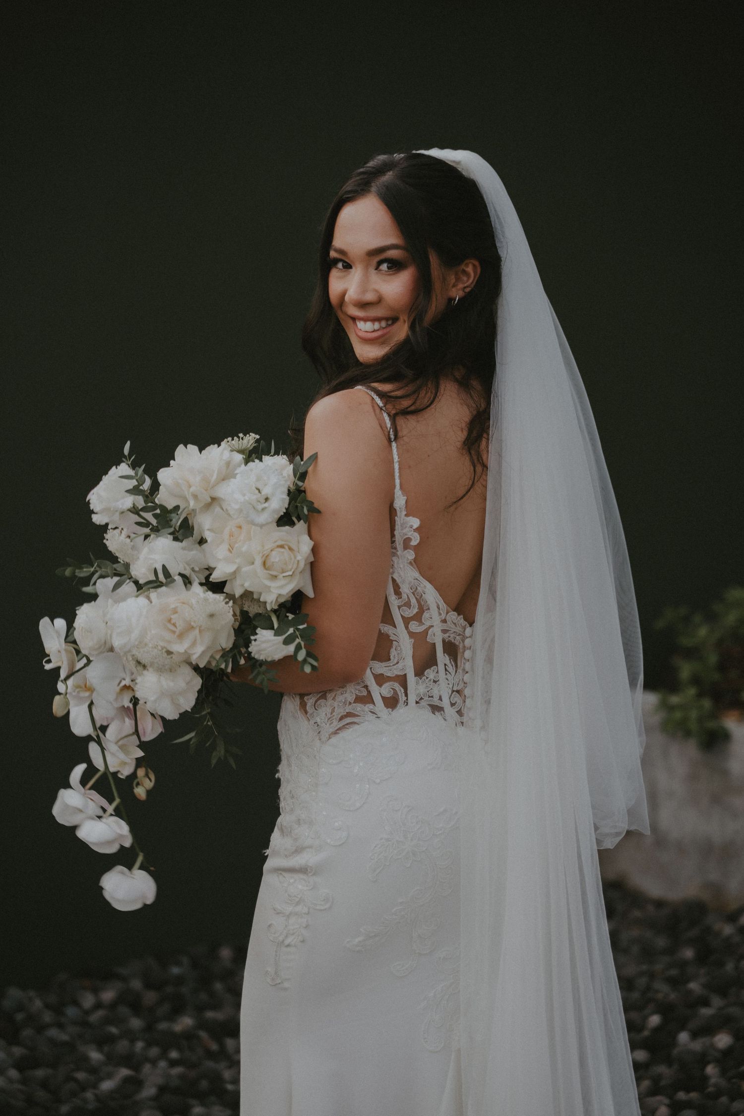 A bride in a white lace wedding dress holds a cascading white orchid bouquet against a dark background.
