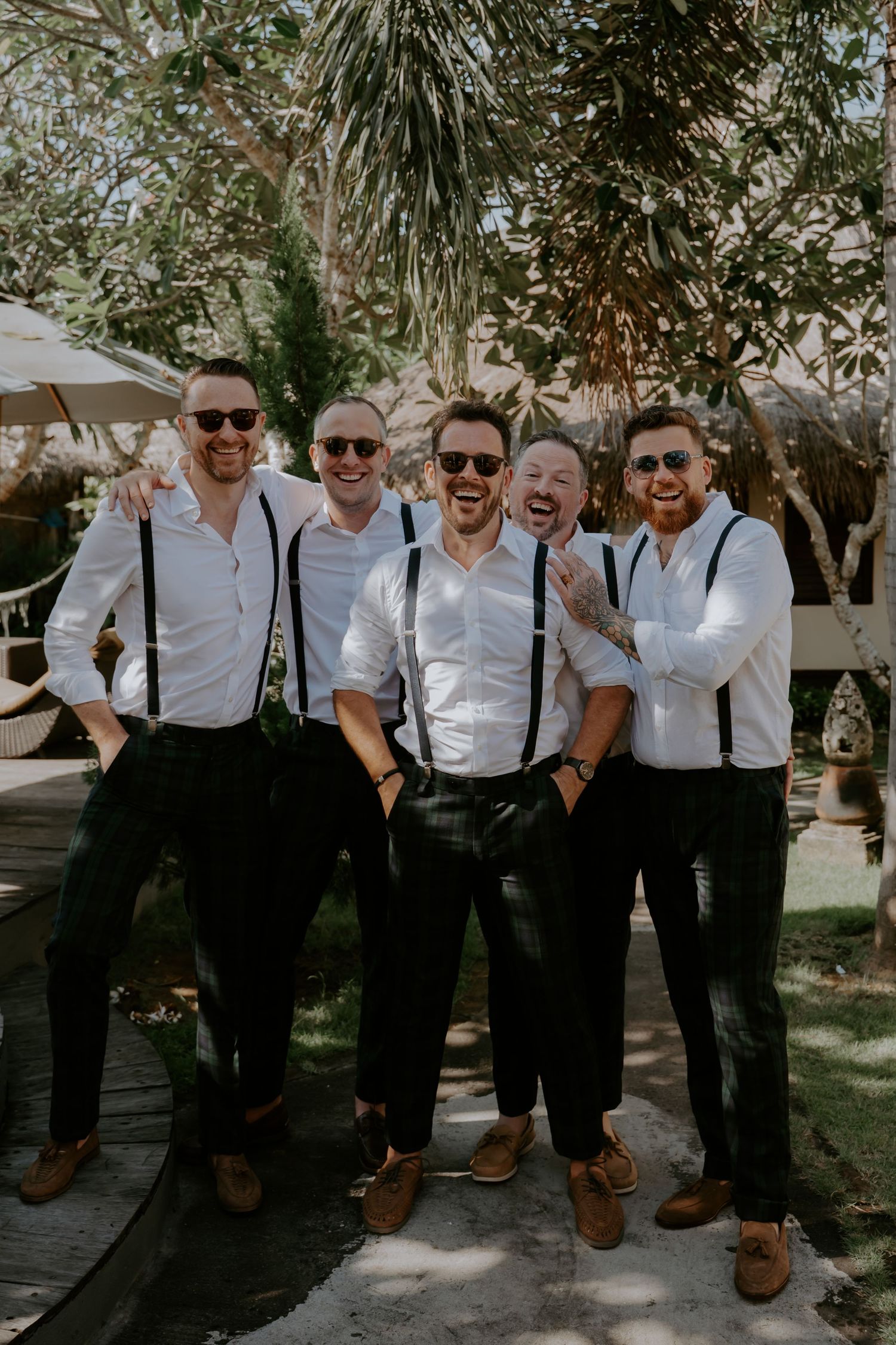 Groomsmen in matching white shirts and black suspenders pose together outdoors for wedding photos.
