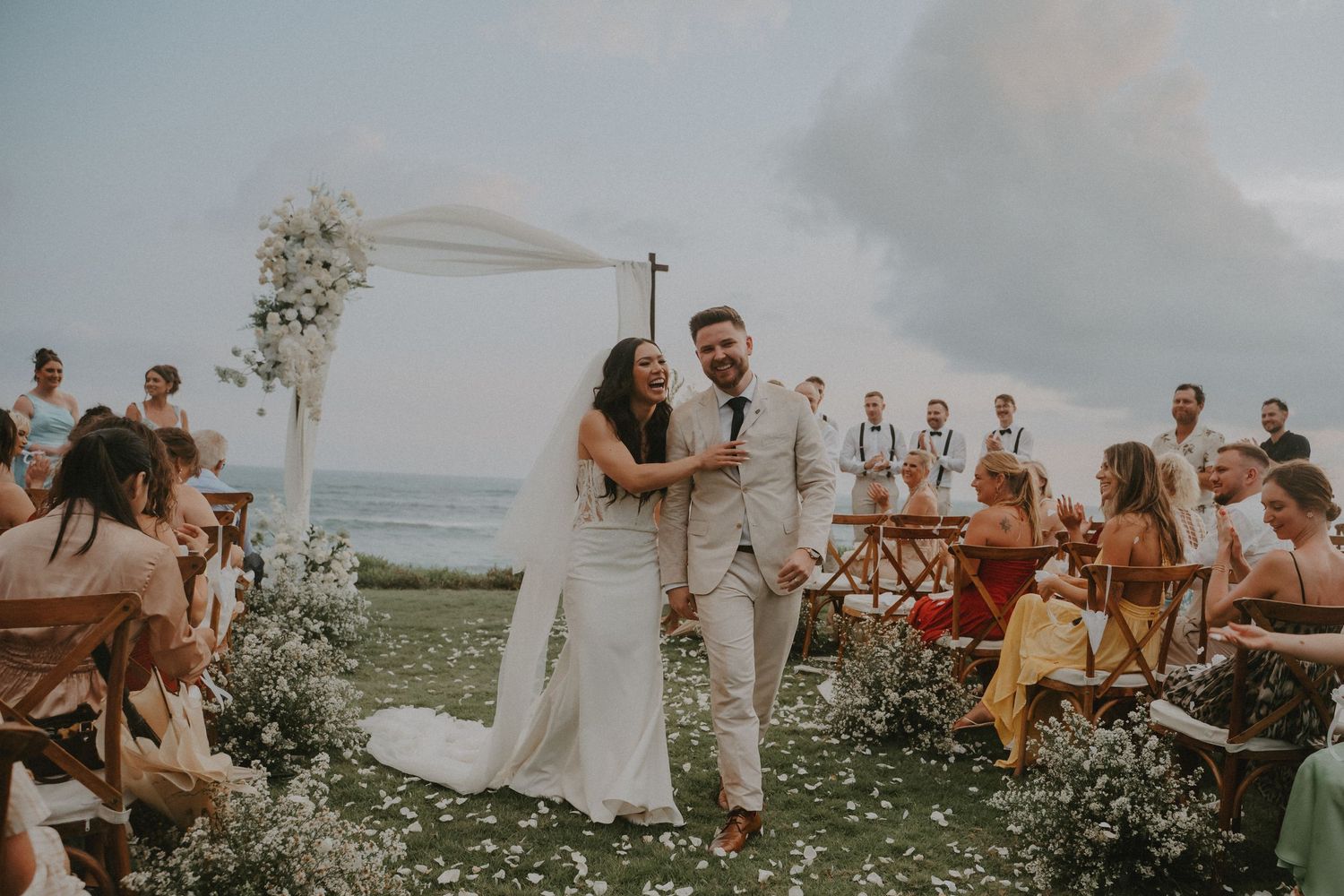 Wedding party celebrates outdoors with white flowers and beige attire against a cloudy sky.
