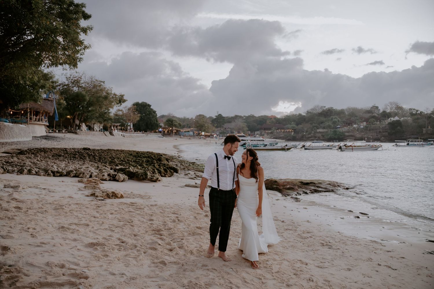 A couple walks along a scenic beach shoreline with cloudy skies and coastal trees in the background.
