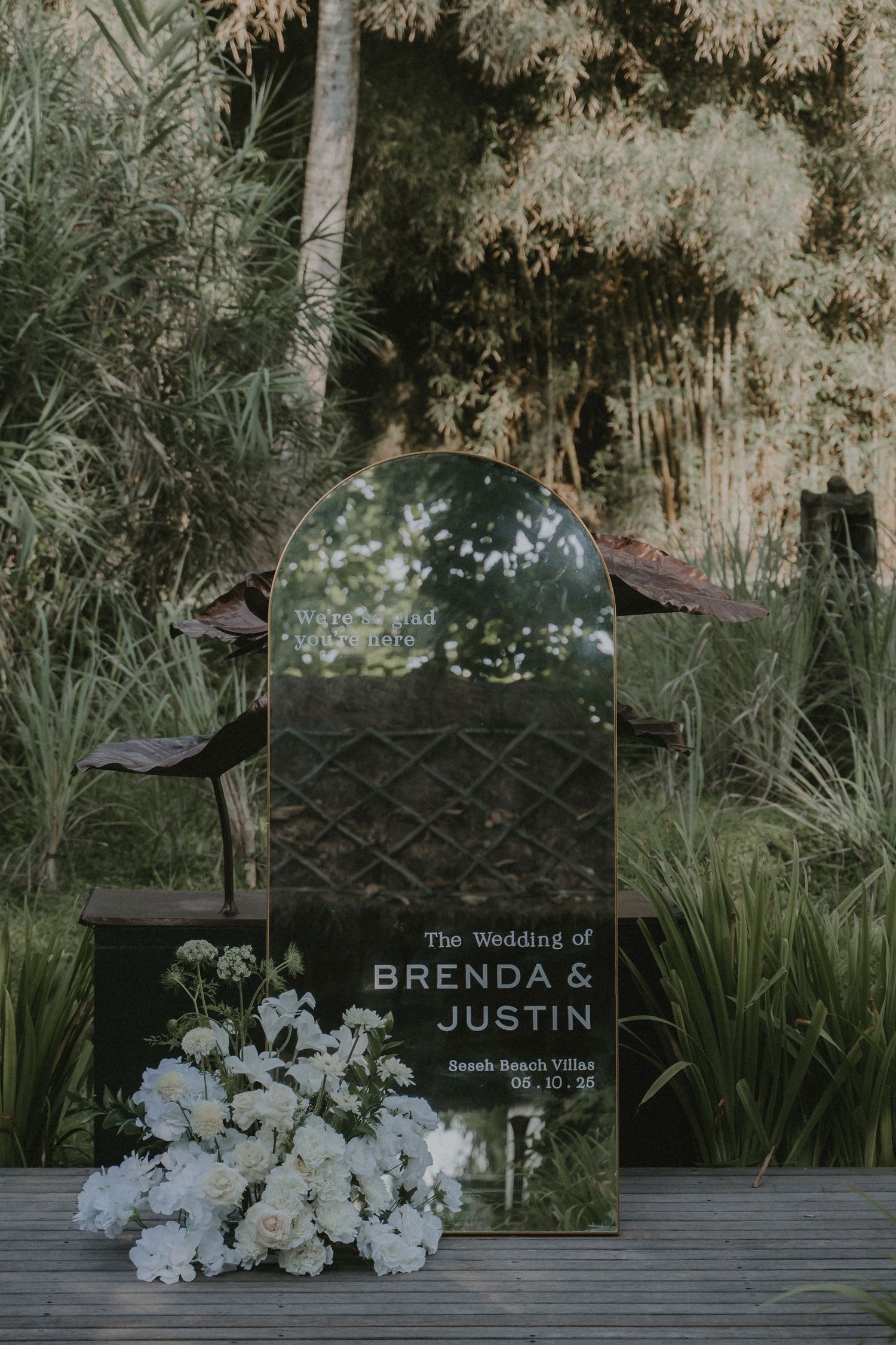 A mirrored wedding sign with white flowers sits in a garden setting surrounded by greenery and bamboo.