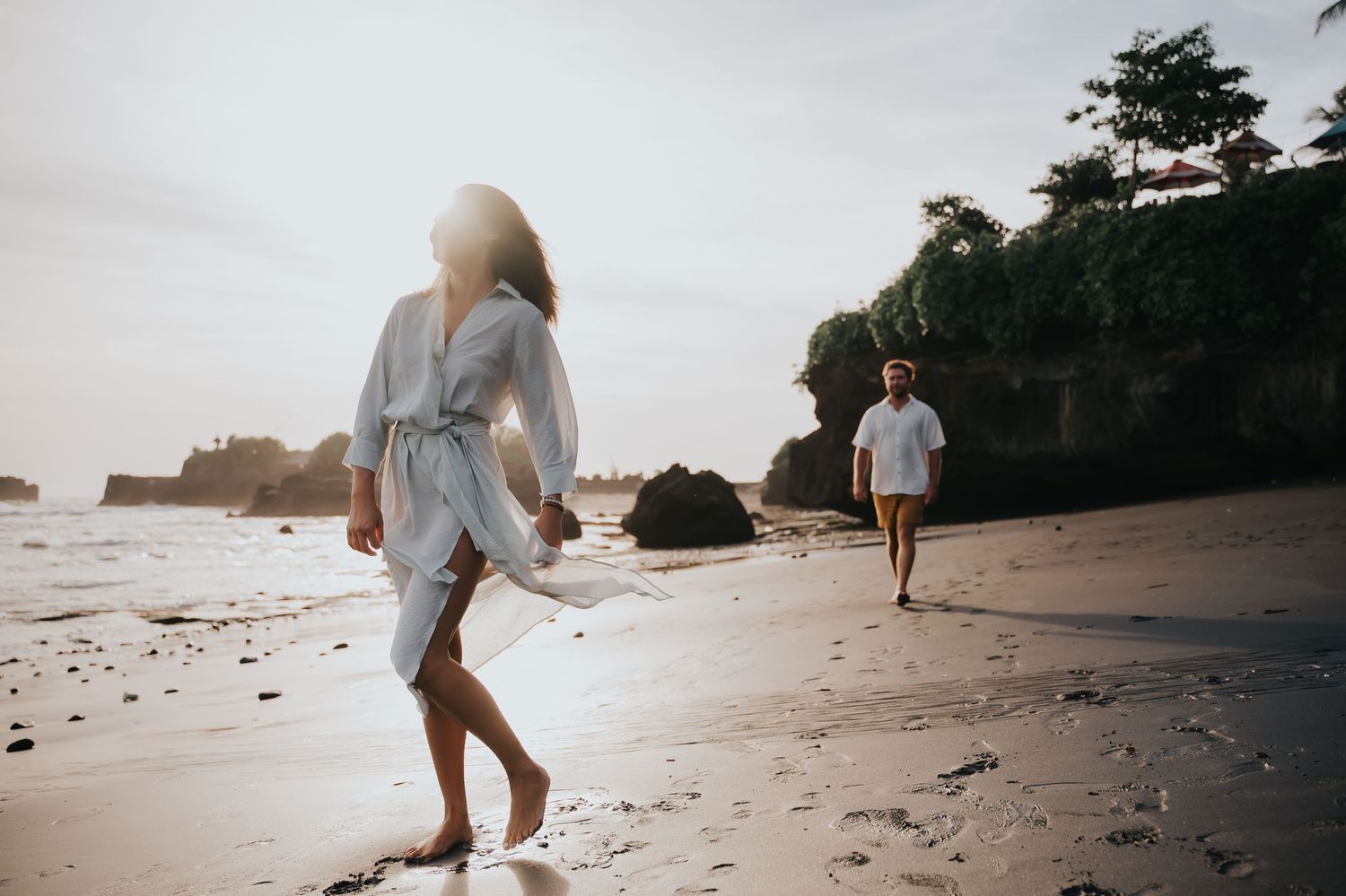 Two people walking along a scenic beach at sunset with rocky cliffs and ocean waves in the background.
