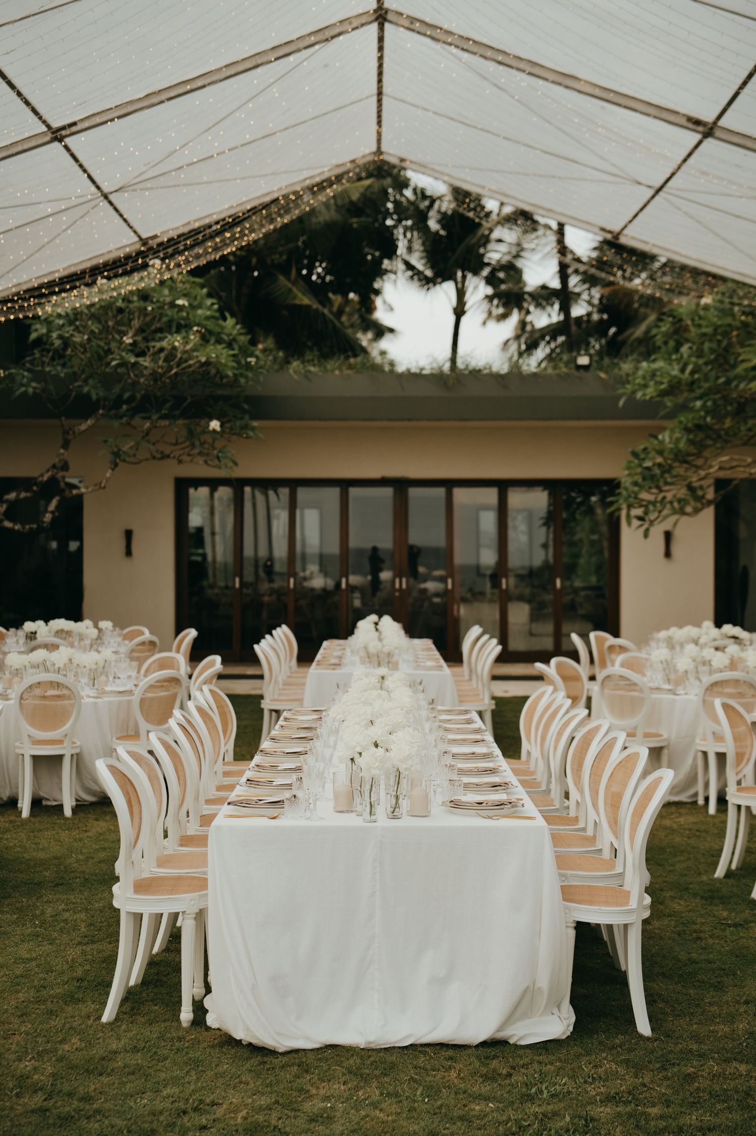 Elegant white wedding reception setup under a clear marquee tent with fairy lights.
