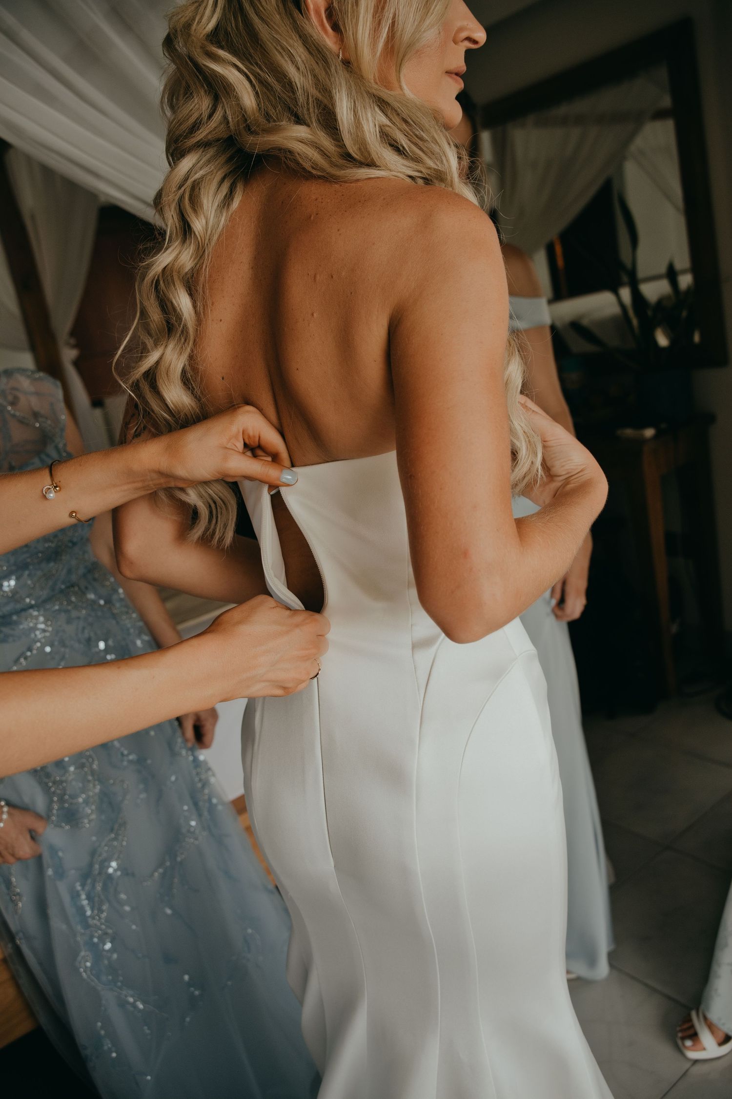 Bridesmaids helping bride lace up her elegant white strapless wedding gown from behind.