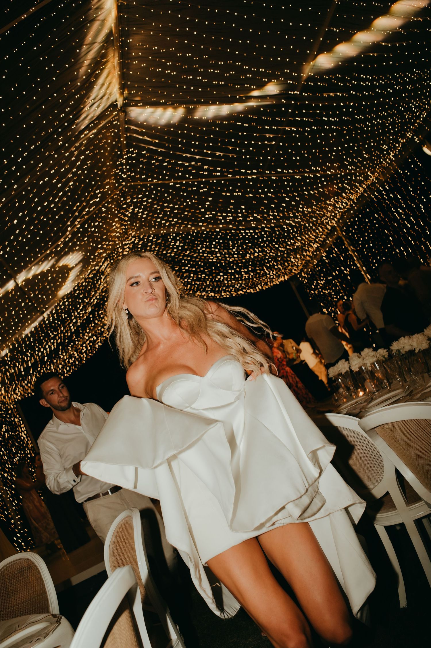 Bride in a short white wedding dress twirls under a stunning canopy of golden fairy lights at her reception.