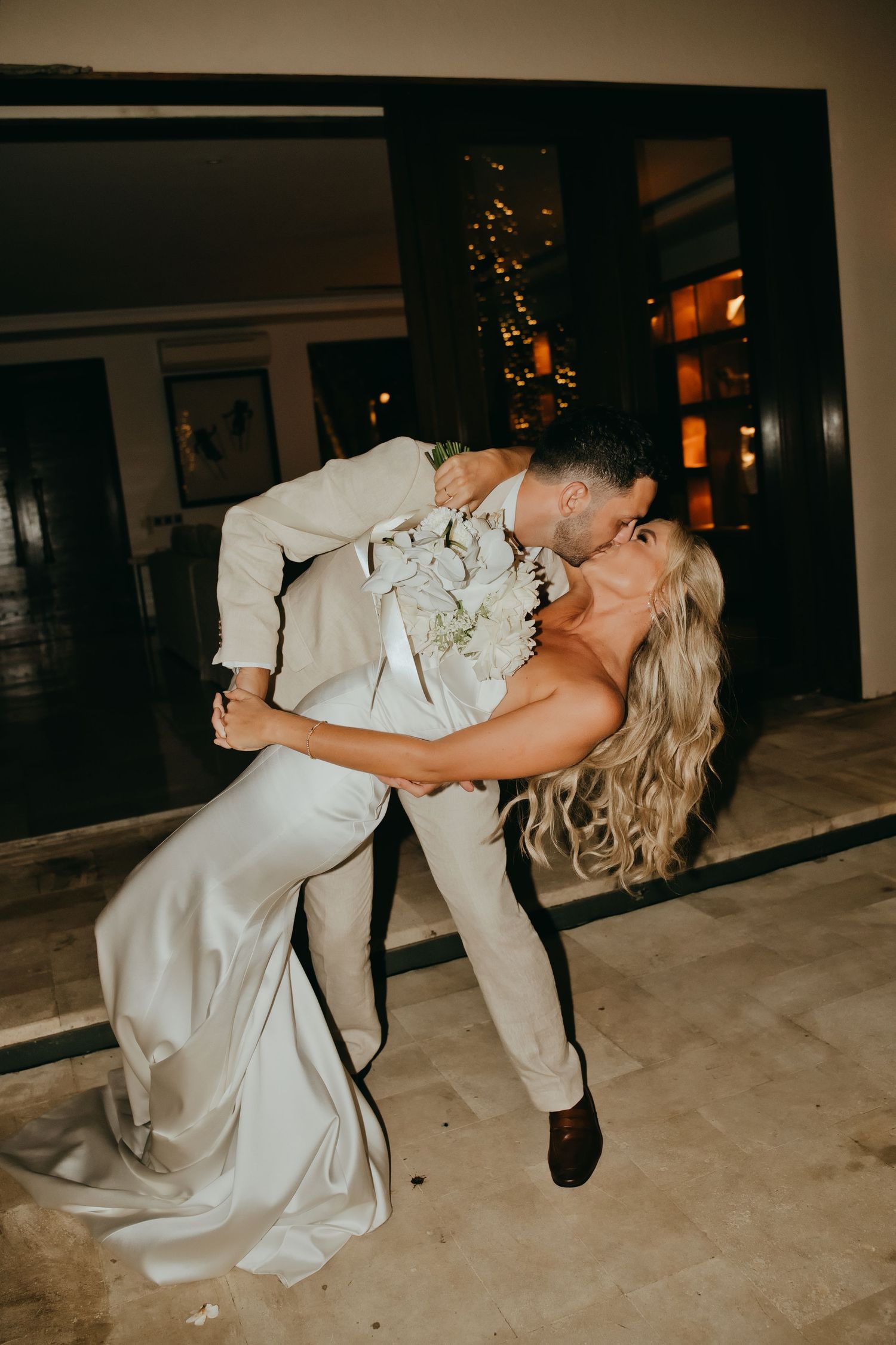 Groom dips bride for a passionate kiss during their outdoor wedding reception at night.
