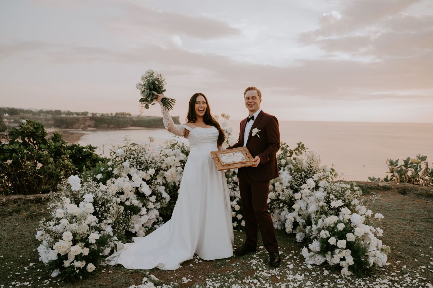 Bride and groom pose with bouquet and wedding certificate at a cliffside ceremony with ocean views.