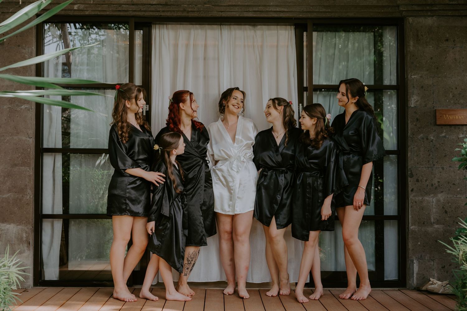 Bride in white robe with six bridesmaids in black satin robes laughing together before wedding ceremony.